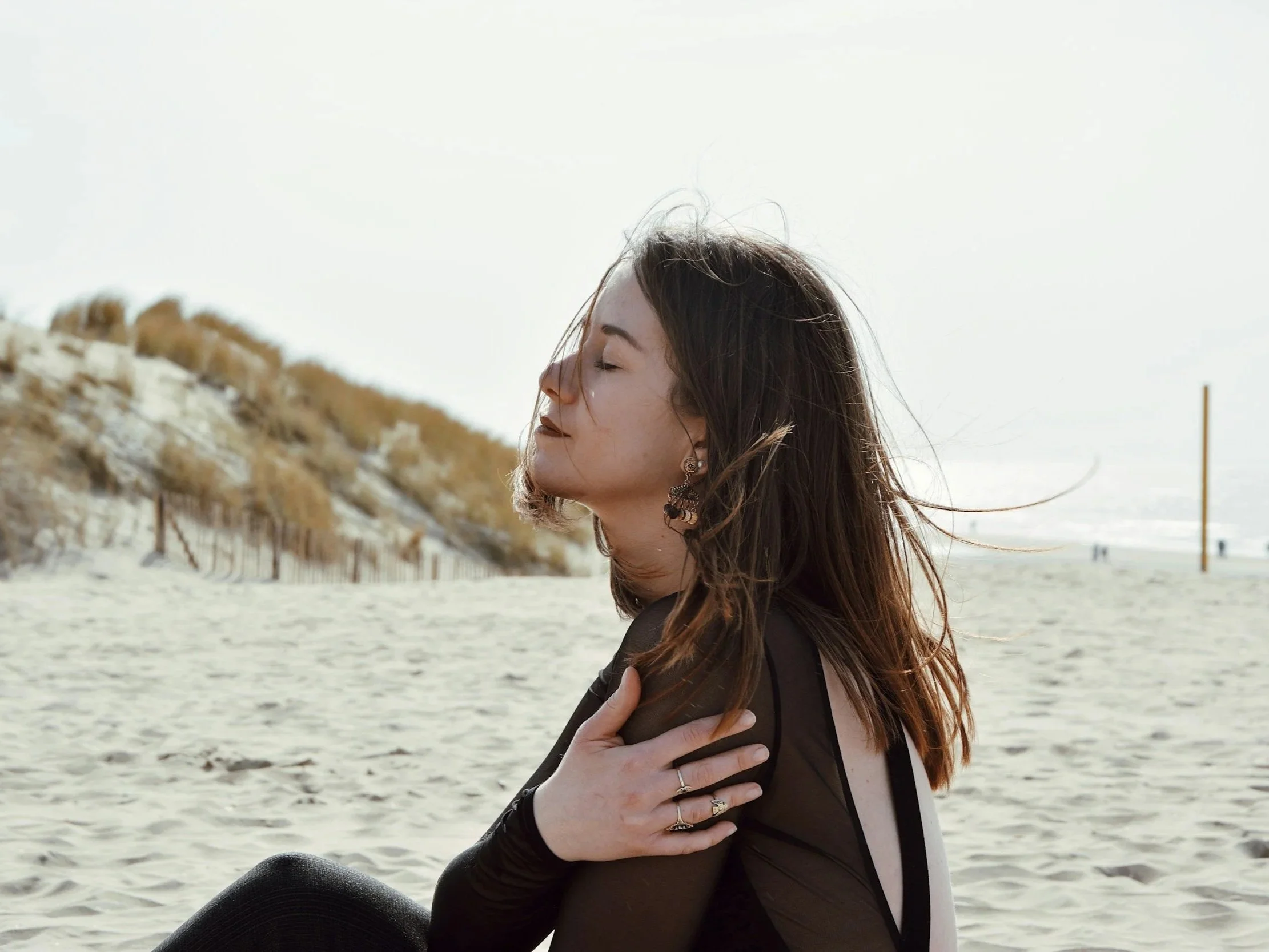 Woman sitting on a beach with eyes closed and hand on her chest, appearing grounded and connected to her body