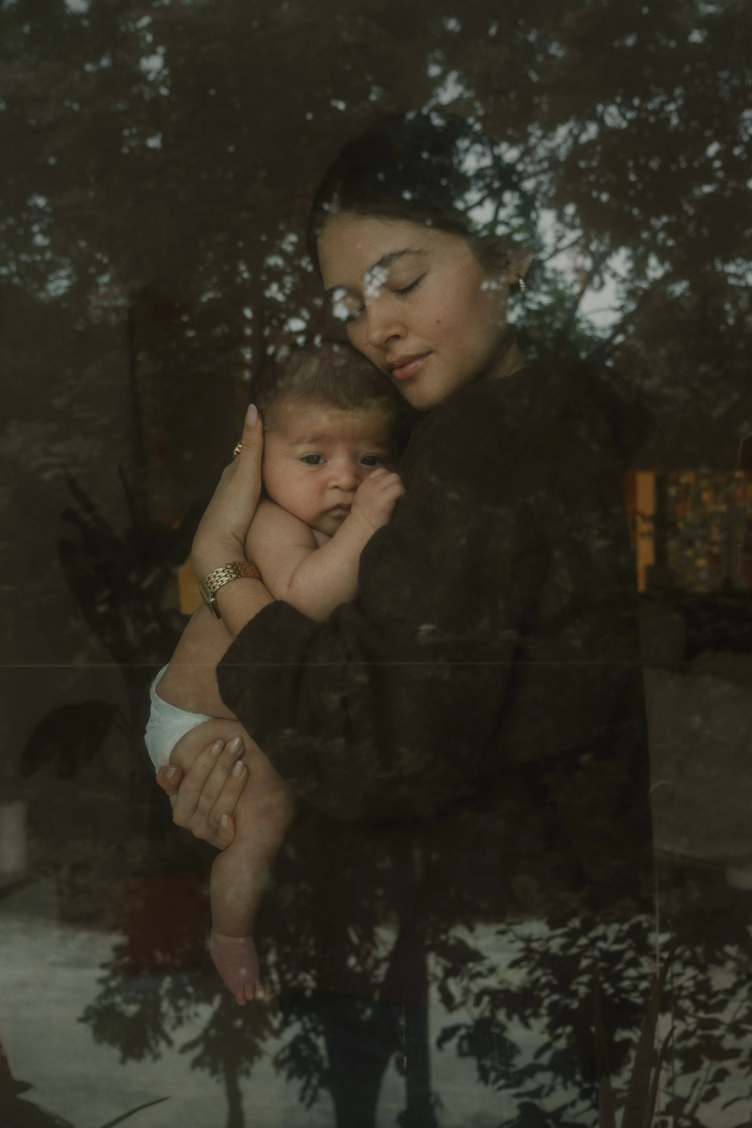 Photo of a woman and her baby behind a window, reflections of trees on the window, captured by Emmedelmar in Cozumel.