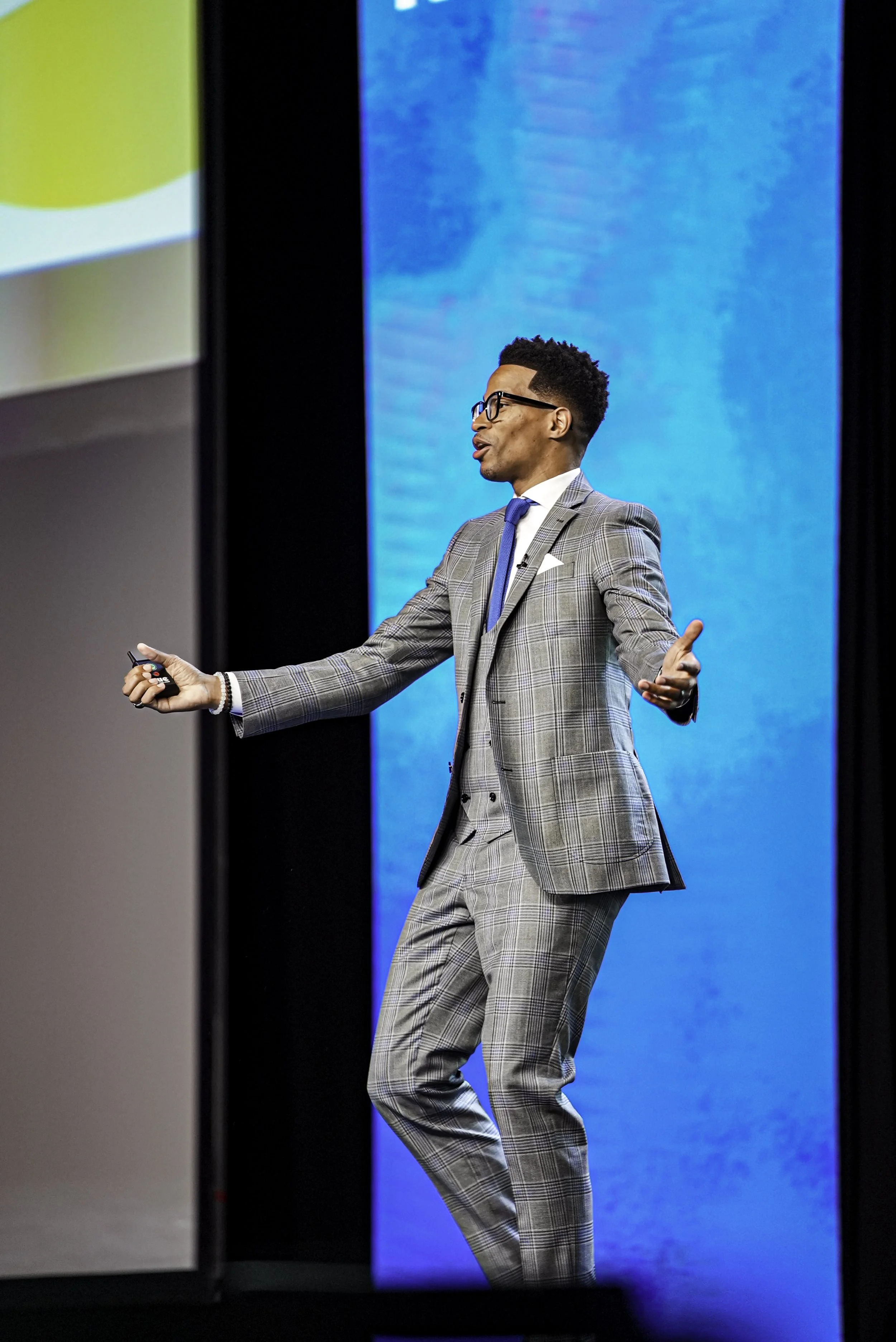 A man wearing glasses, a gray plaid suit, a white shirt, and a blue tie, standing on stage with a blue background, gesturing with both hands during a presentation.