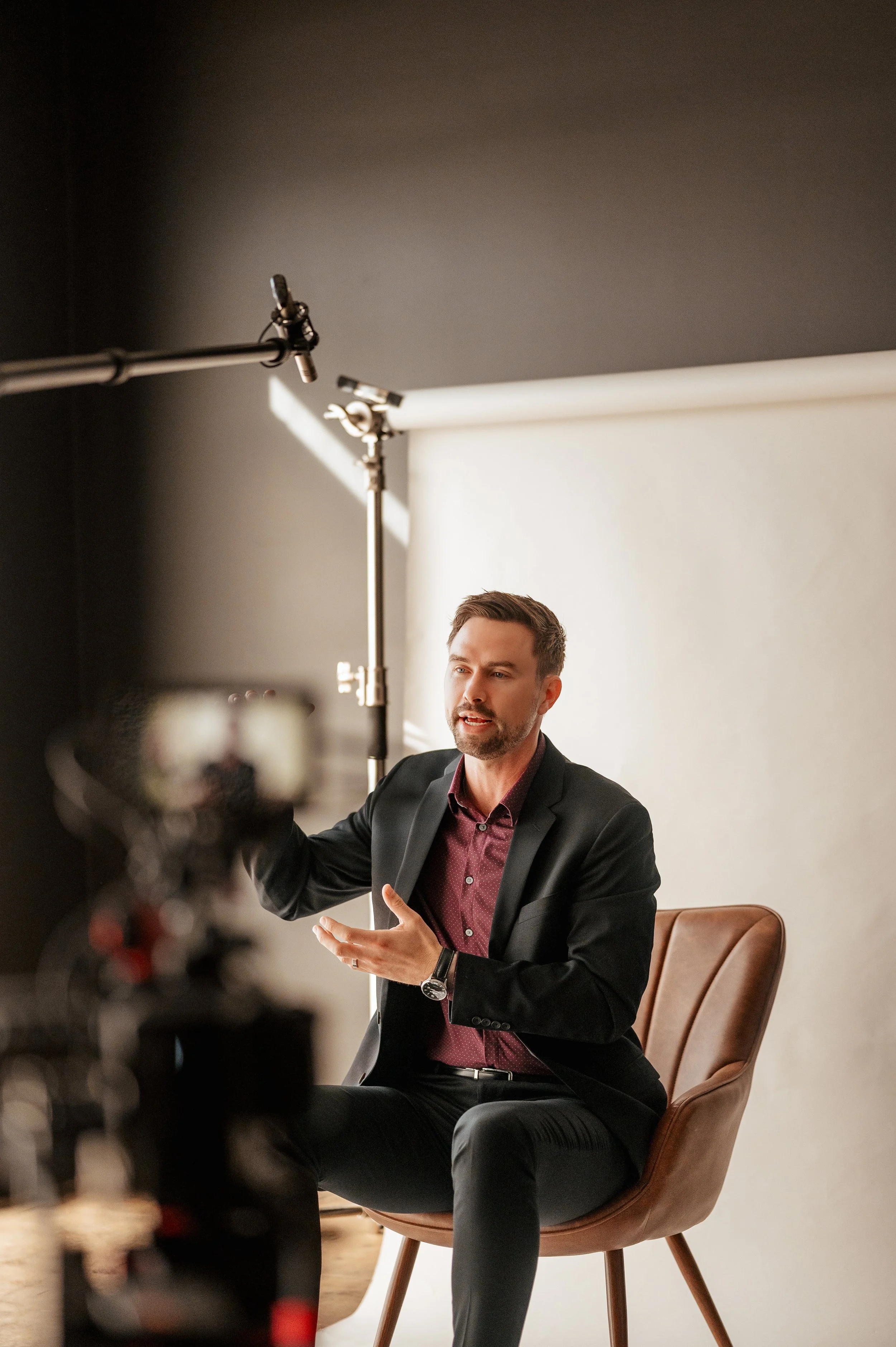 A man in a suit sitting on a brown chair speaking during an interview or video shoot, with professional lighting and equipment visible.