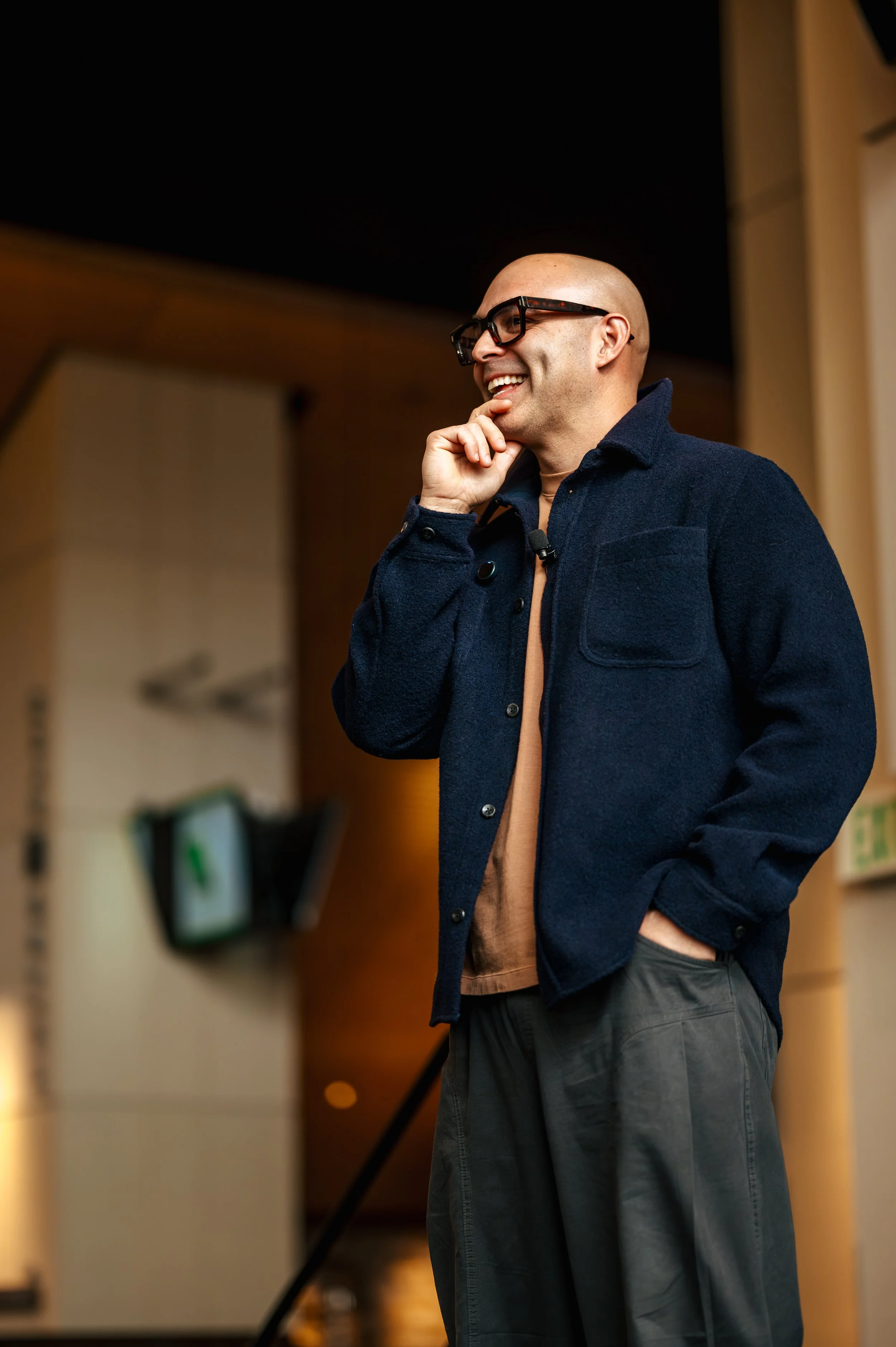 A man with glasses and a bald head, smiling and touching his chin, standing indoors with a dark background and some blurred objects.