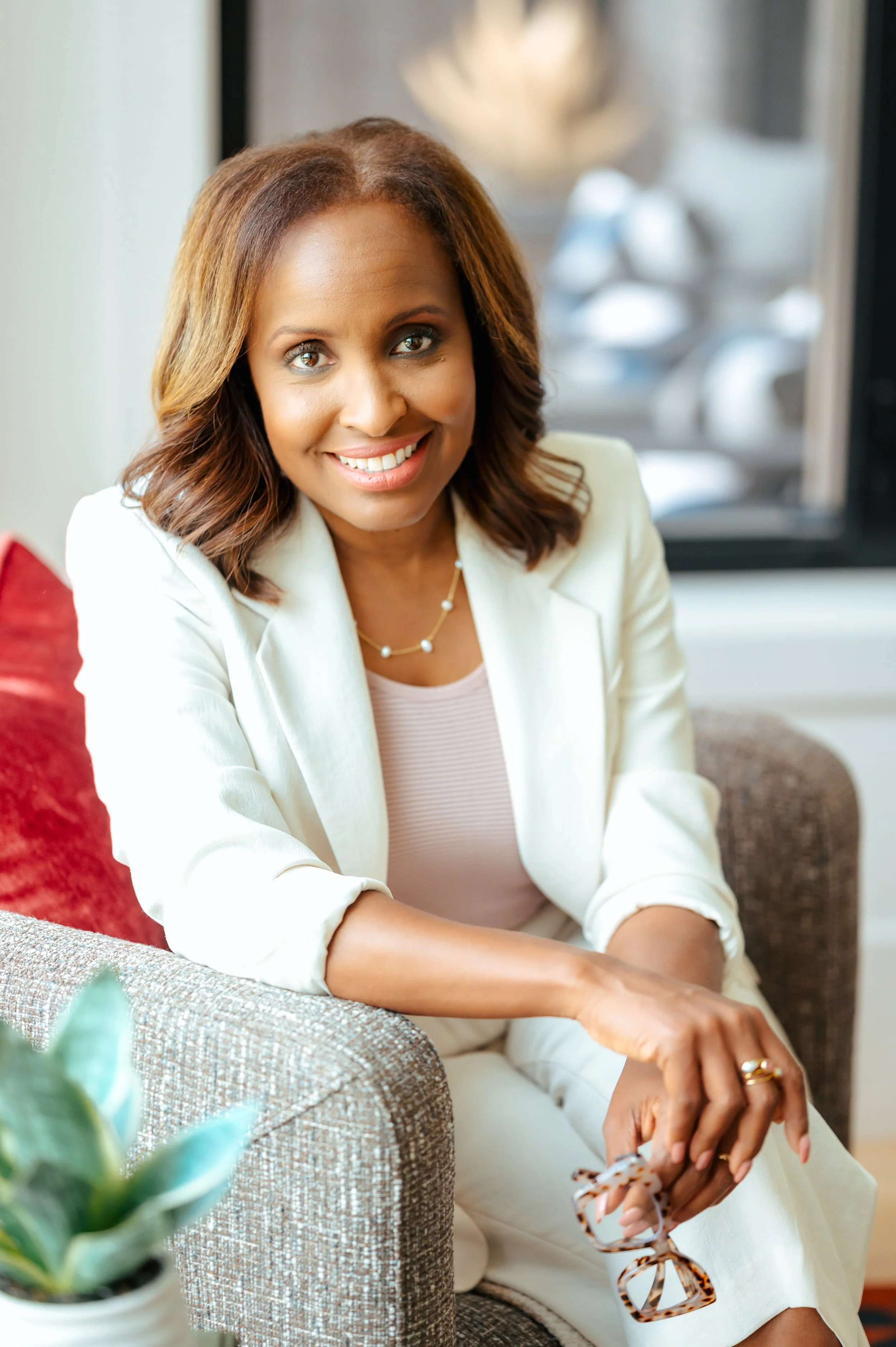 A smiling woman with shoulder-length brown hair seated in a cozy, well-lit room. She wears a light-colored blazer and a pearl necklace, holding a pair of tortoiseshell glasses in her right hand, with a blurred background featuring decorative objects and modern furniture.