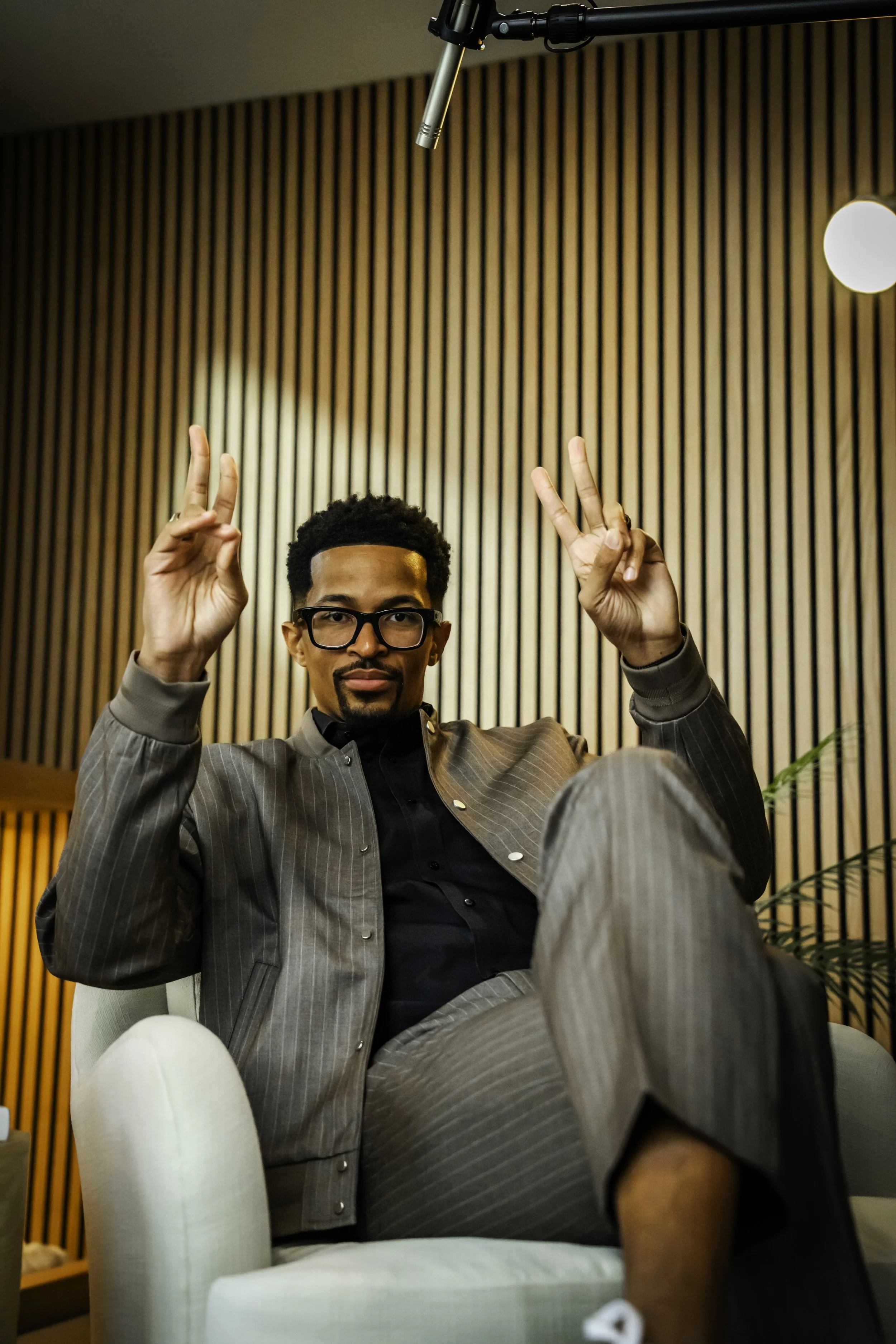 A man with glasses and a beard sitting on a white chair, holding up both peace signs, in a room with wooden vertical paneling on the wall, soft lighting, and a microphone hanging from the ceiling above his head.