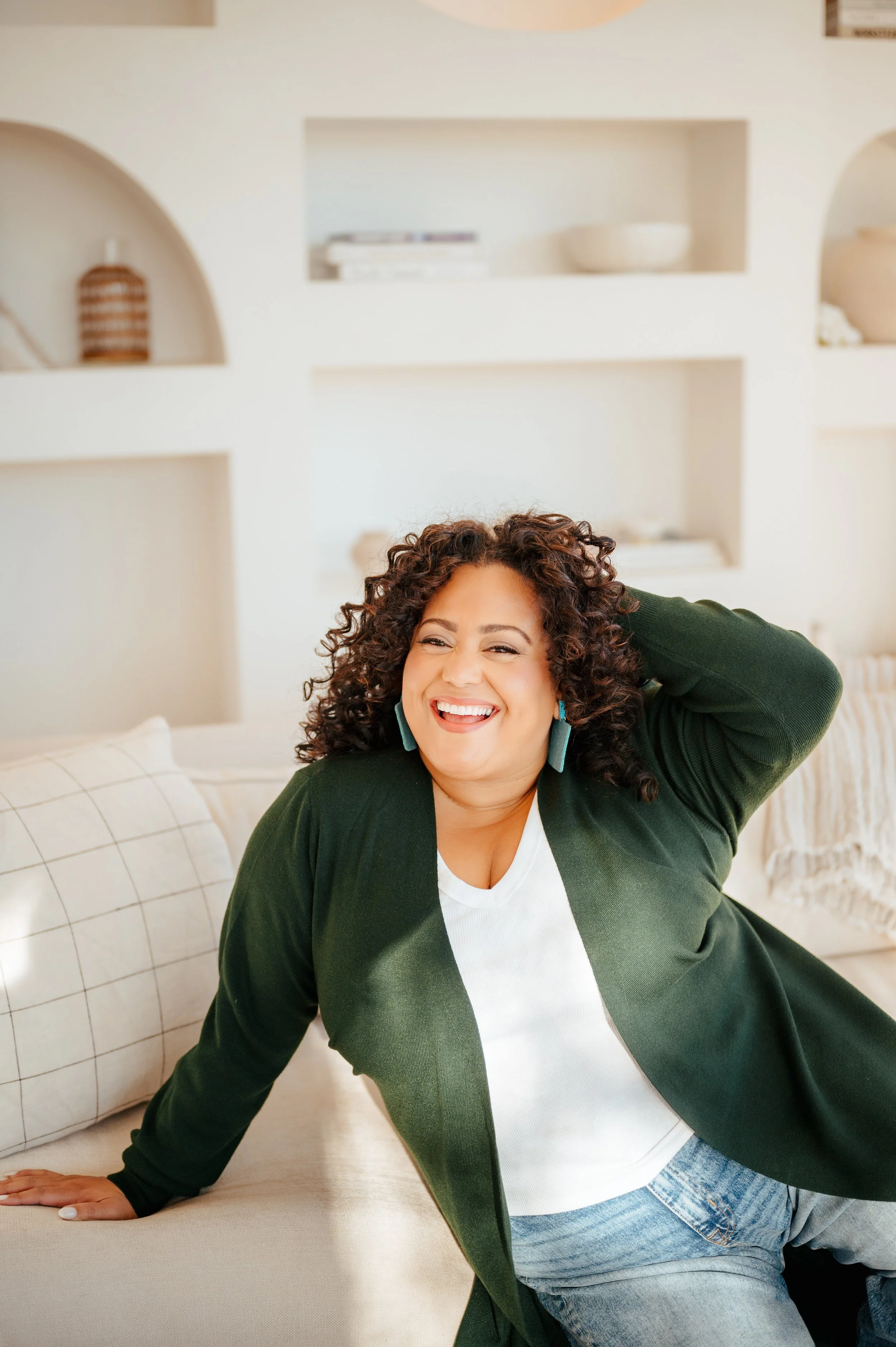 A woman with curly hair smiling and leaning on a beige couch with her hand on the surface, wearing a green blazer and white top, in a bright living room with built-in white shelves holding books and decorative objects.