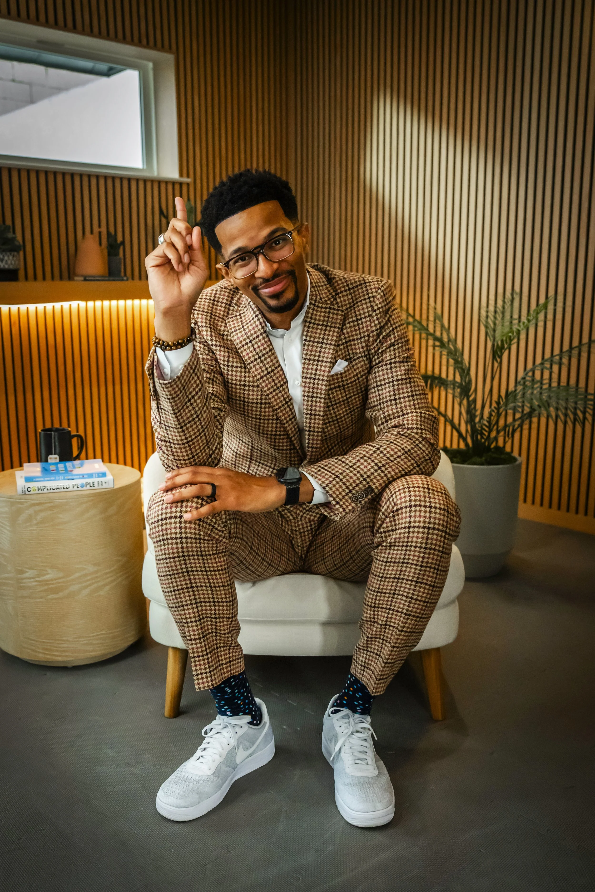 A man in a brown plaid suit and white sneakers sitting on a white chair in a modern room with wooden wall paneling and a tall potted plant. He is smiling, wearing glasses, a black headband, and has a hand raised with one finger pointing up.