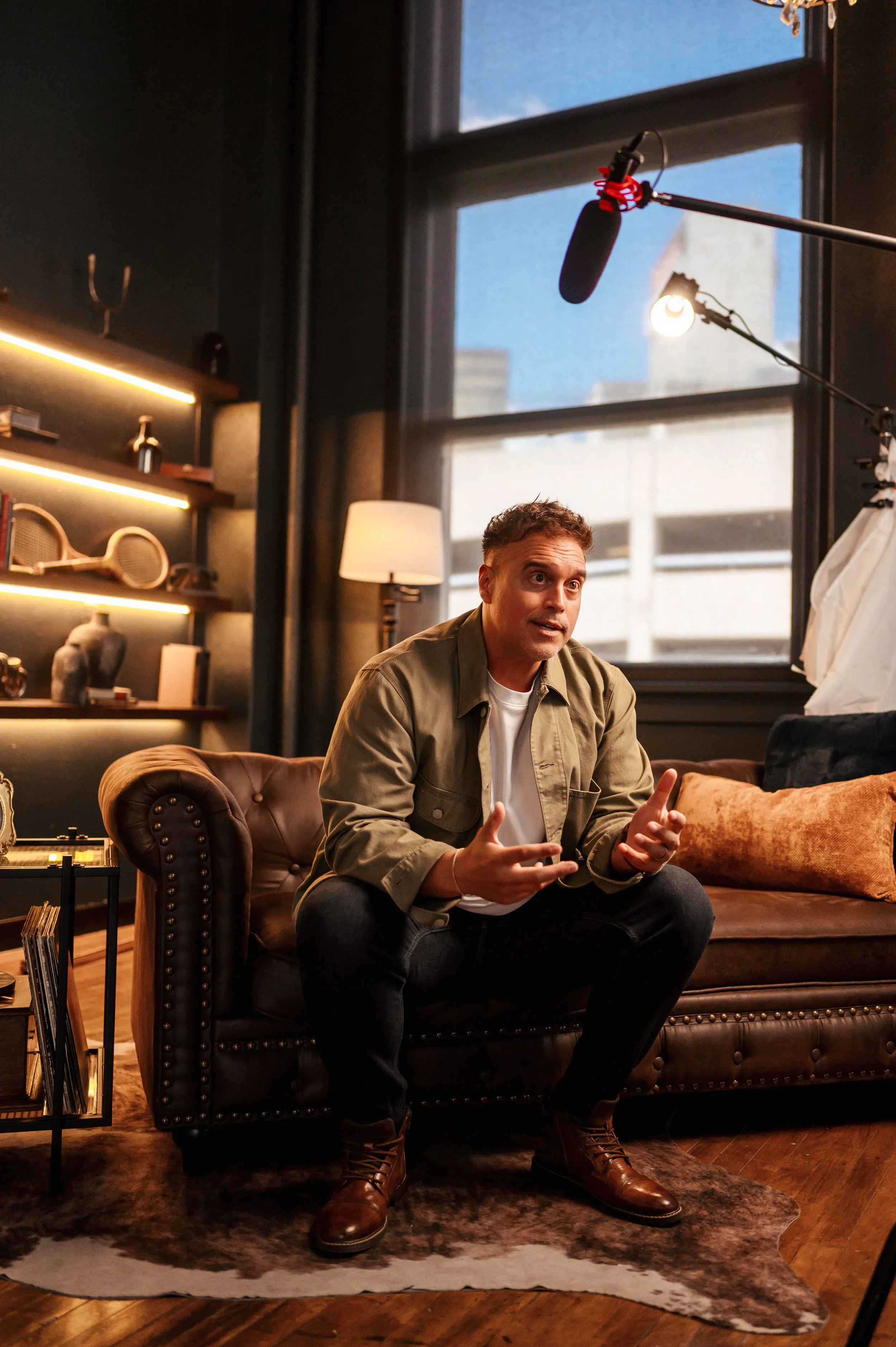 A man sitting on a dark leather couch in a warmly lit room, speaking with hand gestures, with a large window behind him showing buildings and sky.