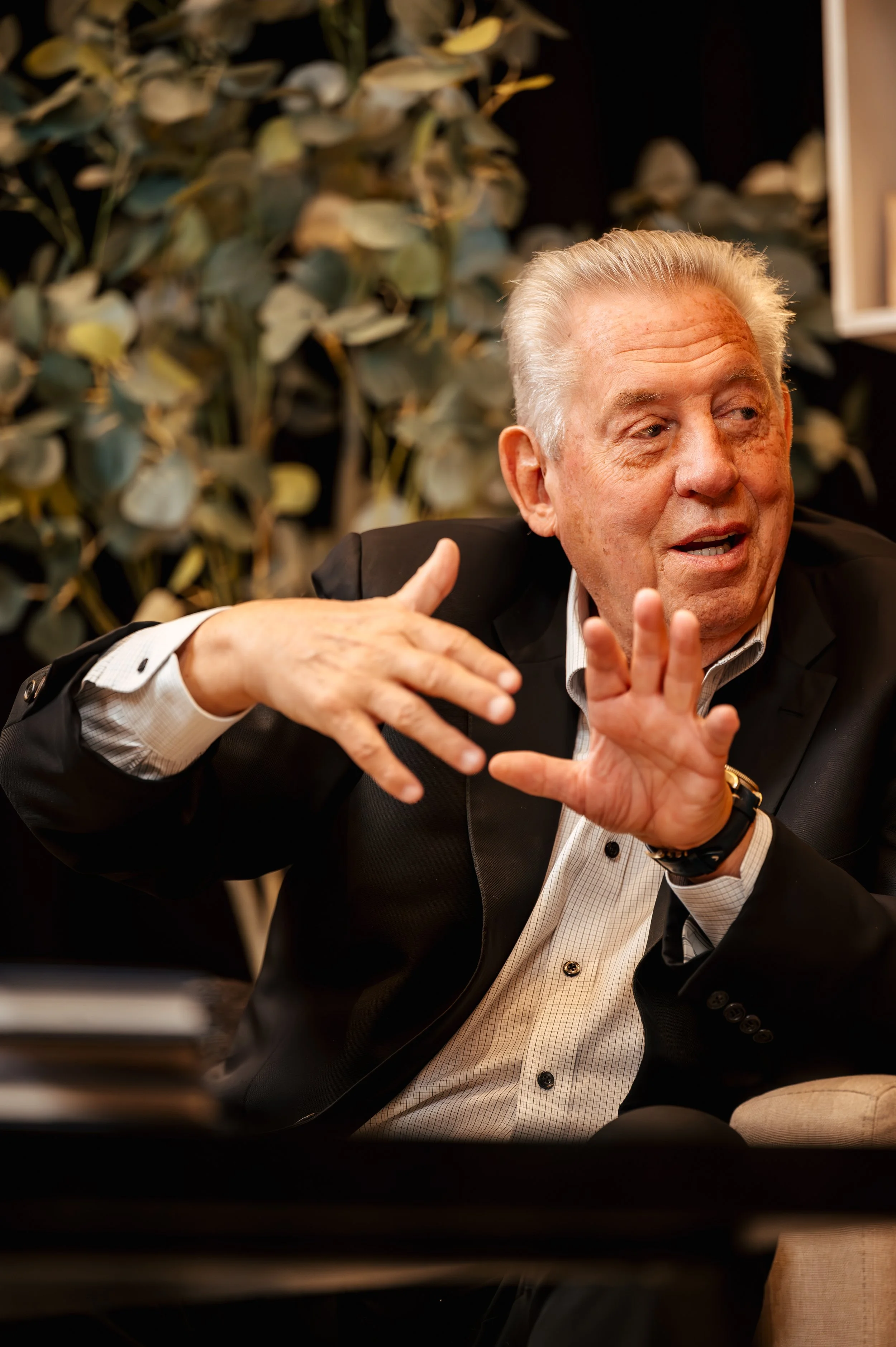 Older man with gray hair, wearing a tuxedo and sitting at a table, gesturing with his hands during a conversation in a dimly lit room with a leafy green plant in the background.