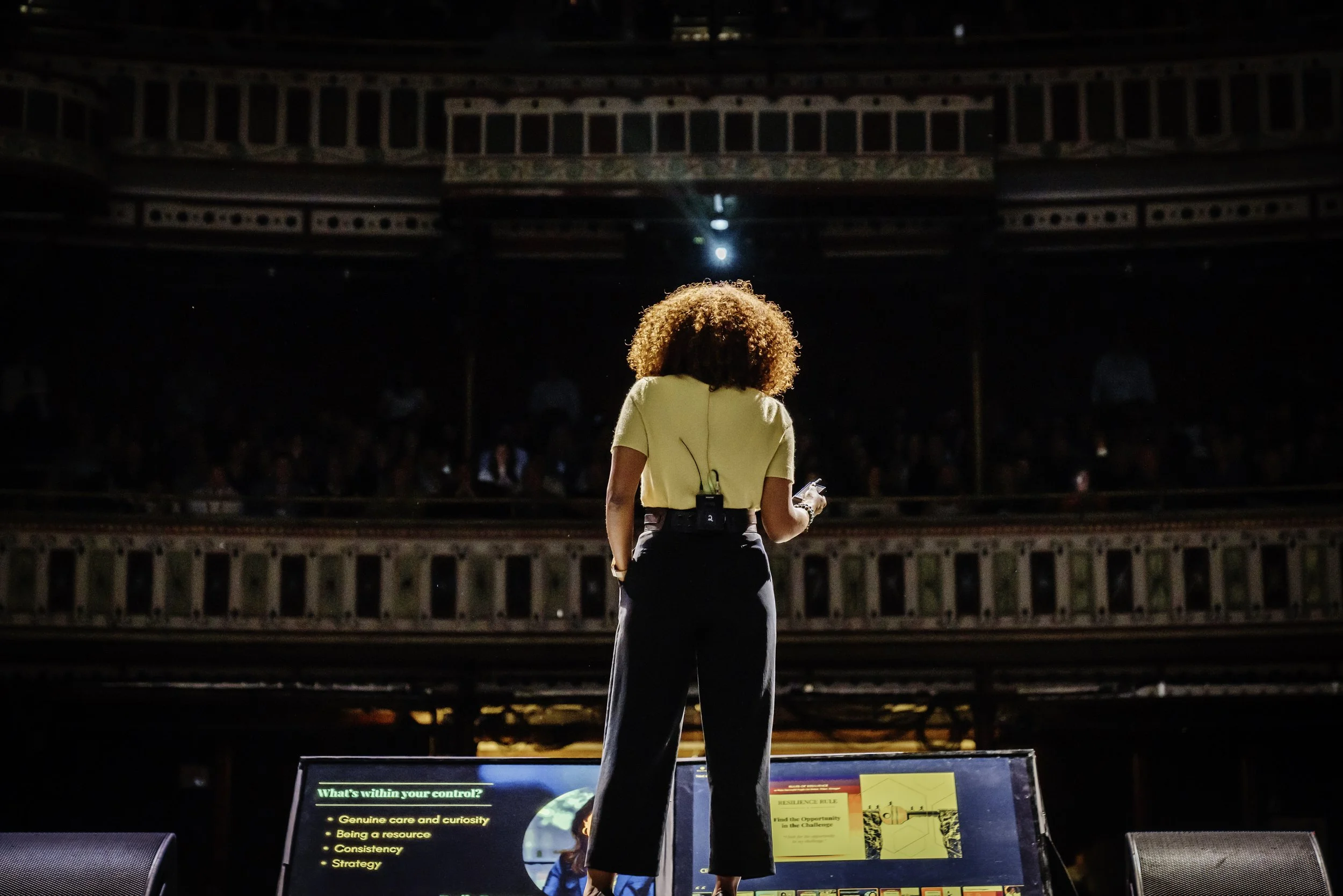 A person with curly hair standing on a stage or platform, facing a large audience in a theater or auditorium. They are looking at a screen displaying presentation slides with text.