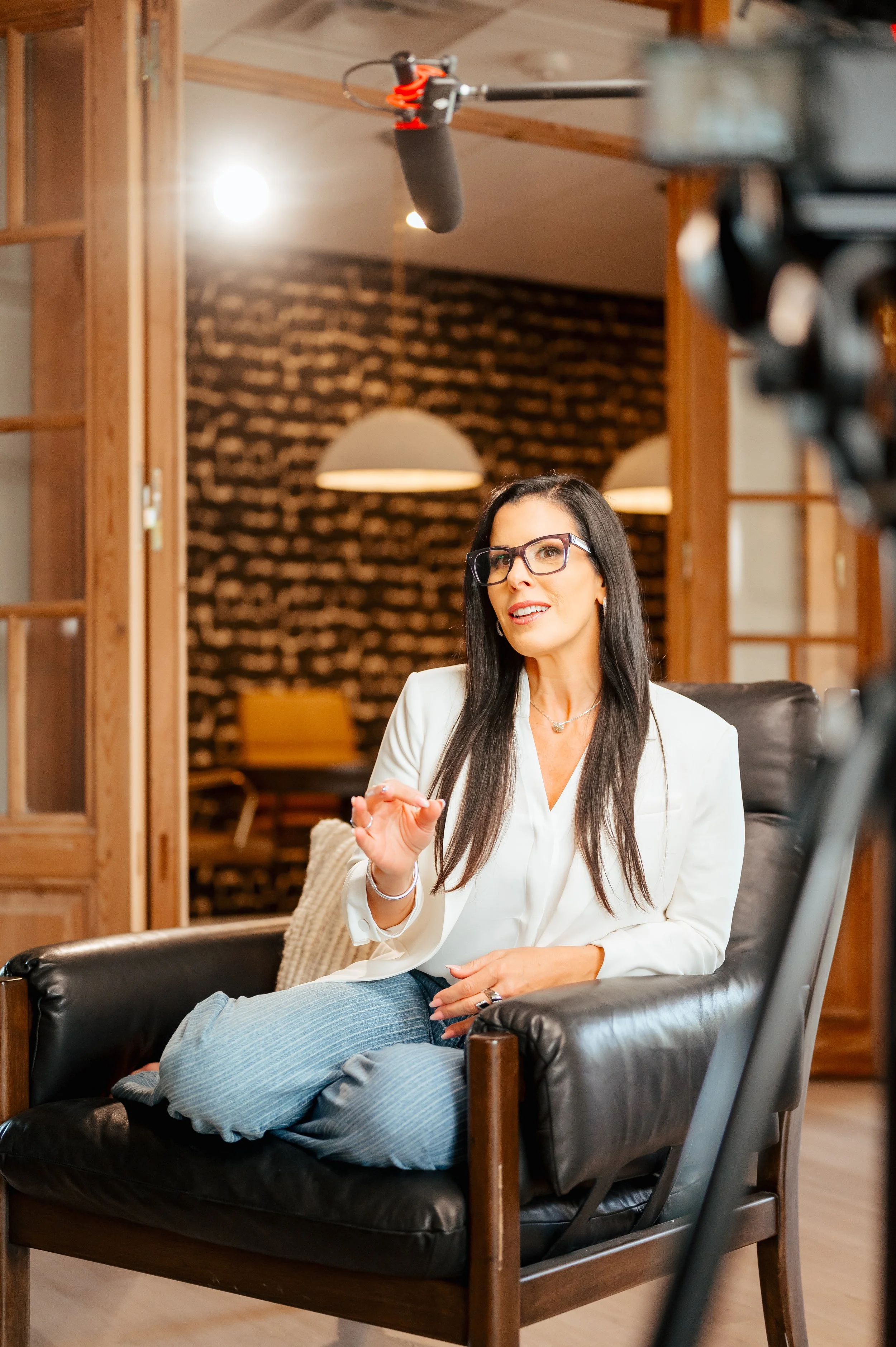 A woman with long dark hair, glasses, and a white blazer sitting on a black leather couch, chatting with a person lying down with legs crossed in a modern, warmly lit room with exposed brick wall background.
