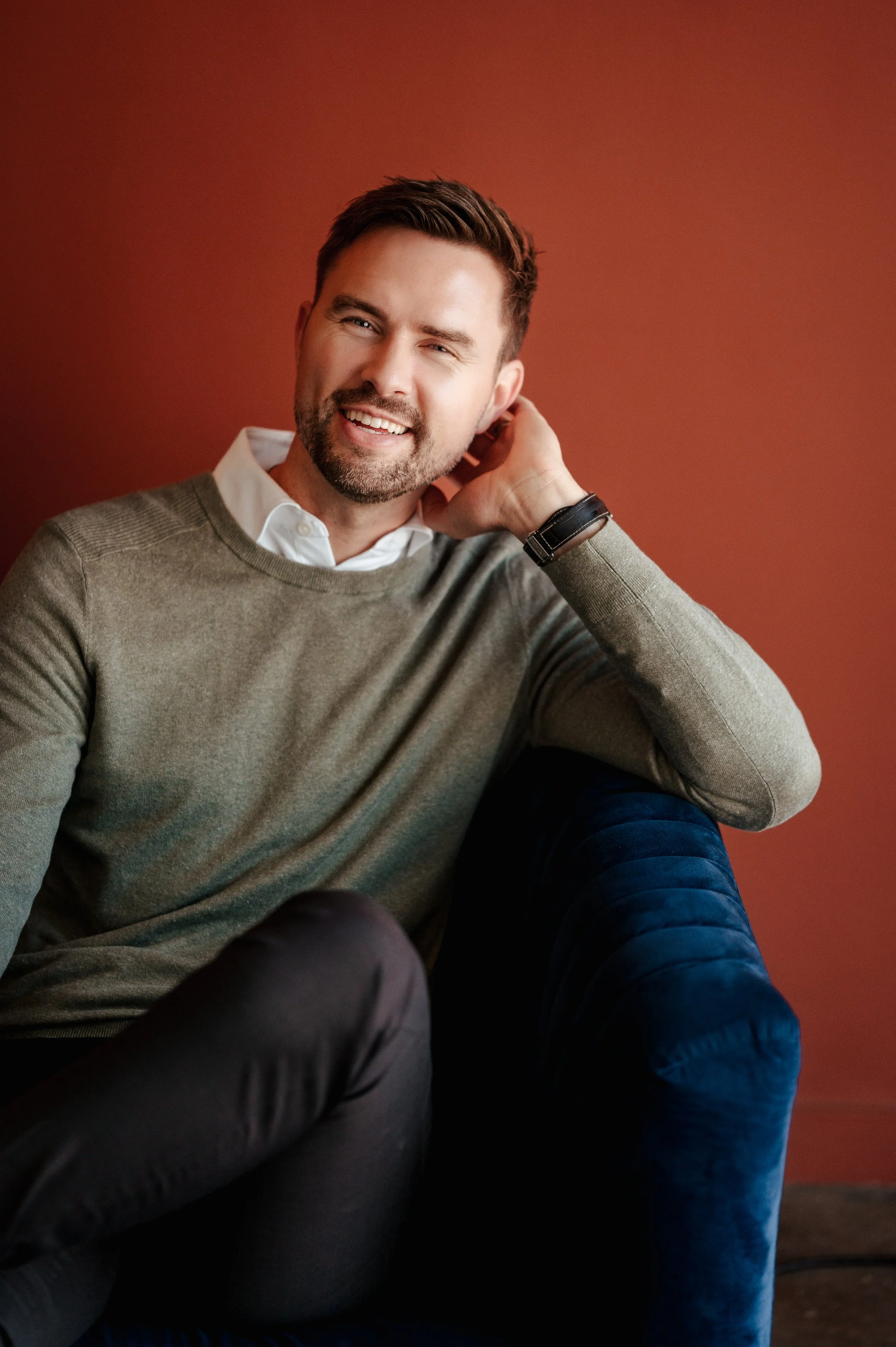 A man with dark hair and a beard, smiling and tilting his head slightly while touching the back of his neck, wearing a white shirt under a beige sweater and a wristwatch, sitting against a reddish-brown background.