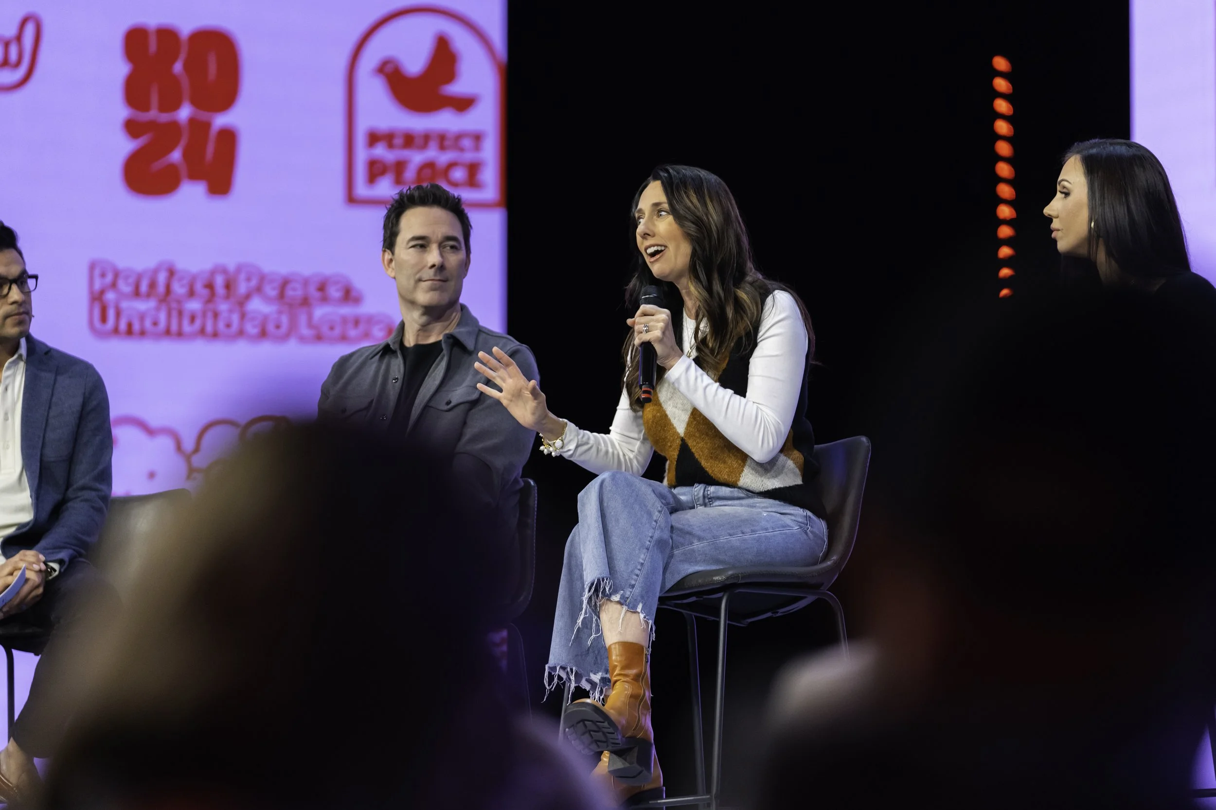 A panel of four people seated on stage during a discussion, with a woman speaking into a microphone. A large screen behind them displays a logo and text. The stage has a dark background with red vertical lights.