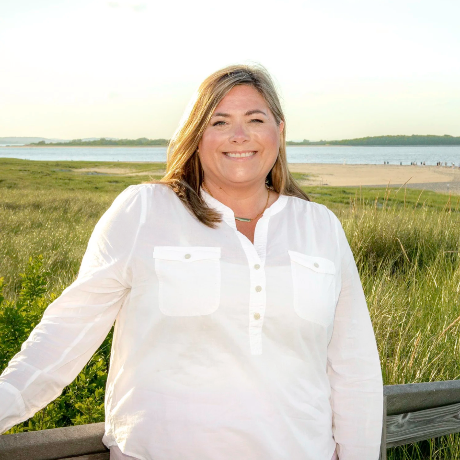 A woman smiling outdoors near a body of water and grassy fields, dressed in a white shirt.