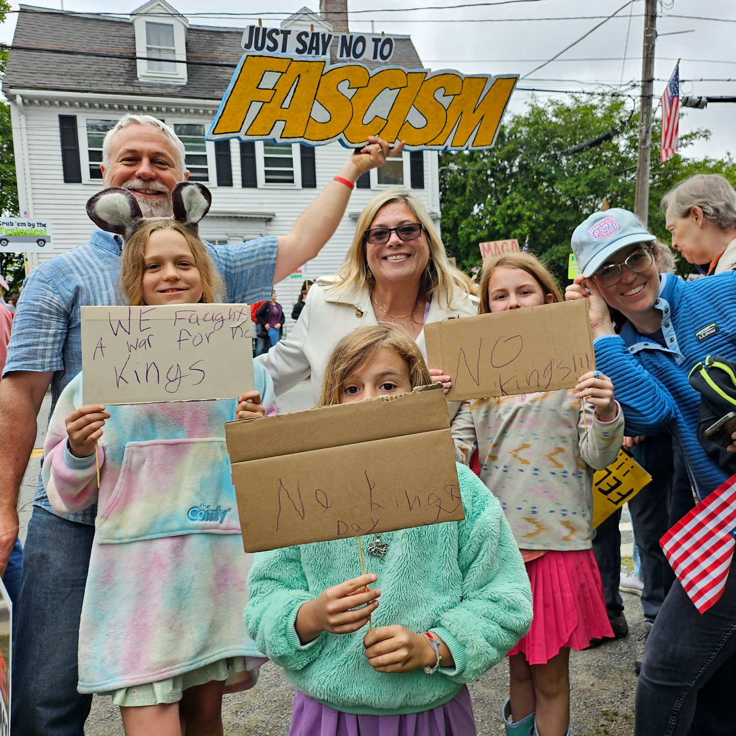 A group of protesters, including children and adults, holding signs that oppose fascism. The signs read 'WE fought a war for the kings' and 'NO kings!!!' Others hold a large sign that says 'Just say no to fascism.' The setting appears to be a street in a residential neighborhood.