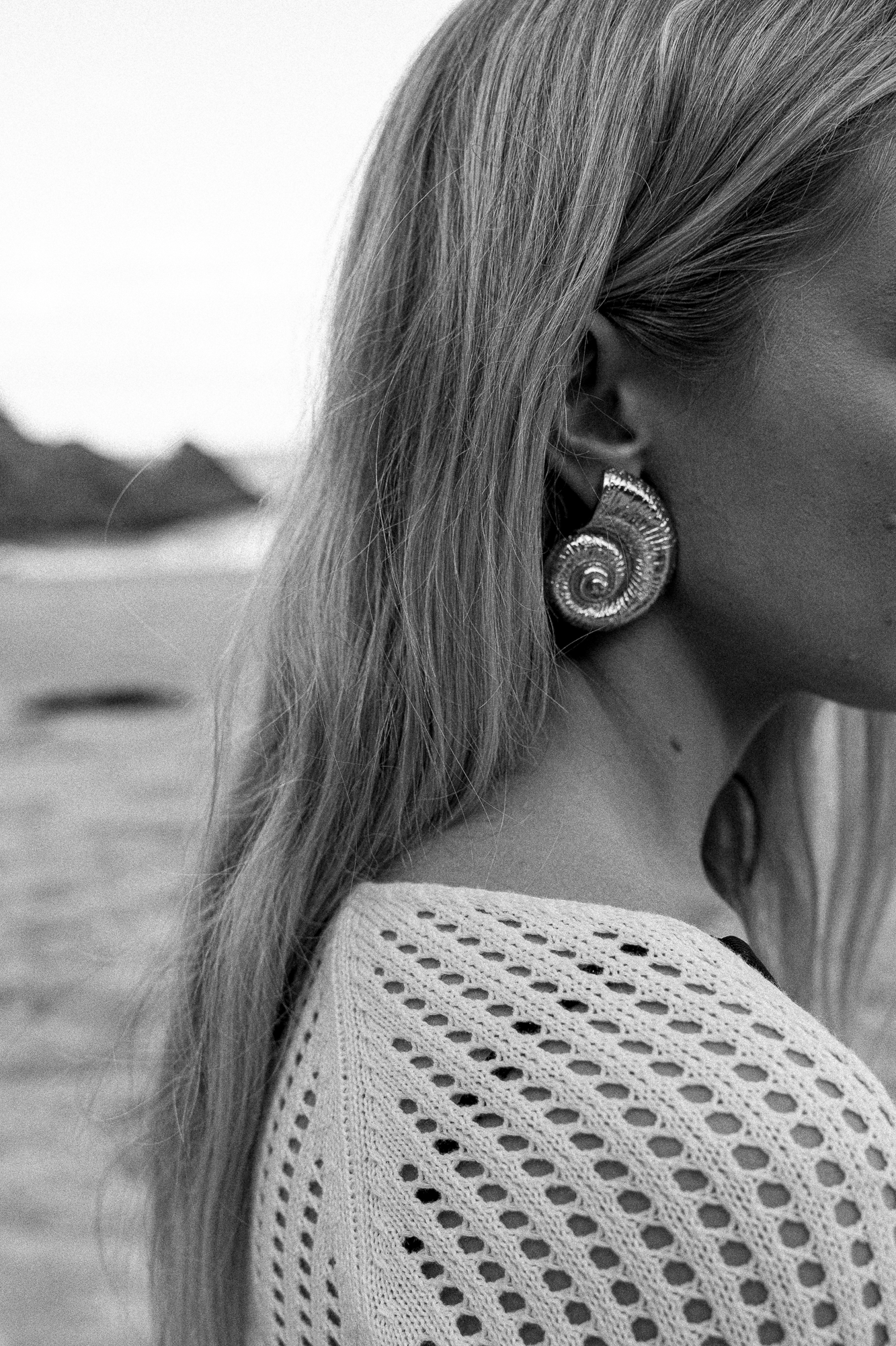 Close-up of a woman with long hair wearing a large, spiral-shaped earring and a knitted, open-weave top, with a blurred beach and distant mountains in the background.