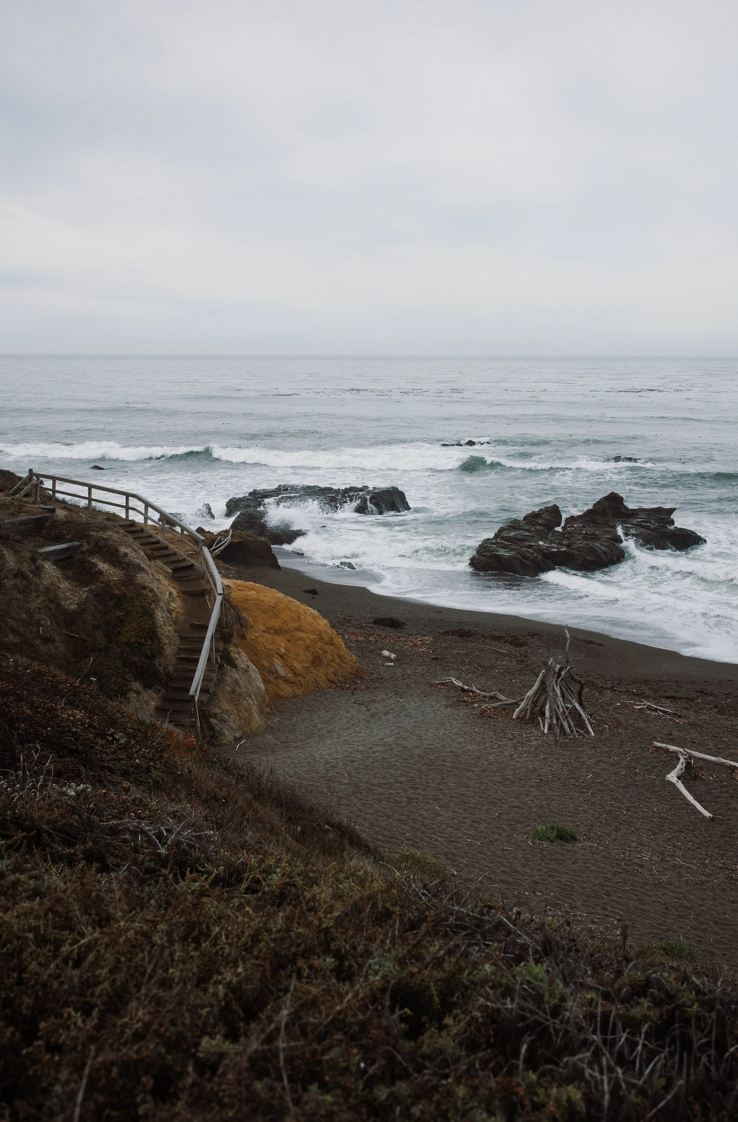 A rocky beach with a staircase leading down from a hillside, driftwood on the sand, and the ocean with waves in the background under an overcast sky.