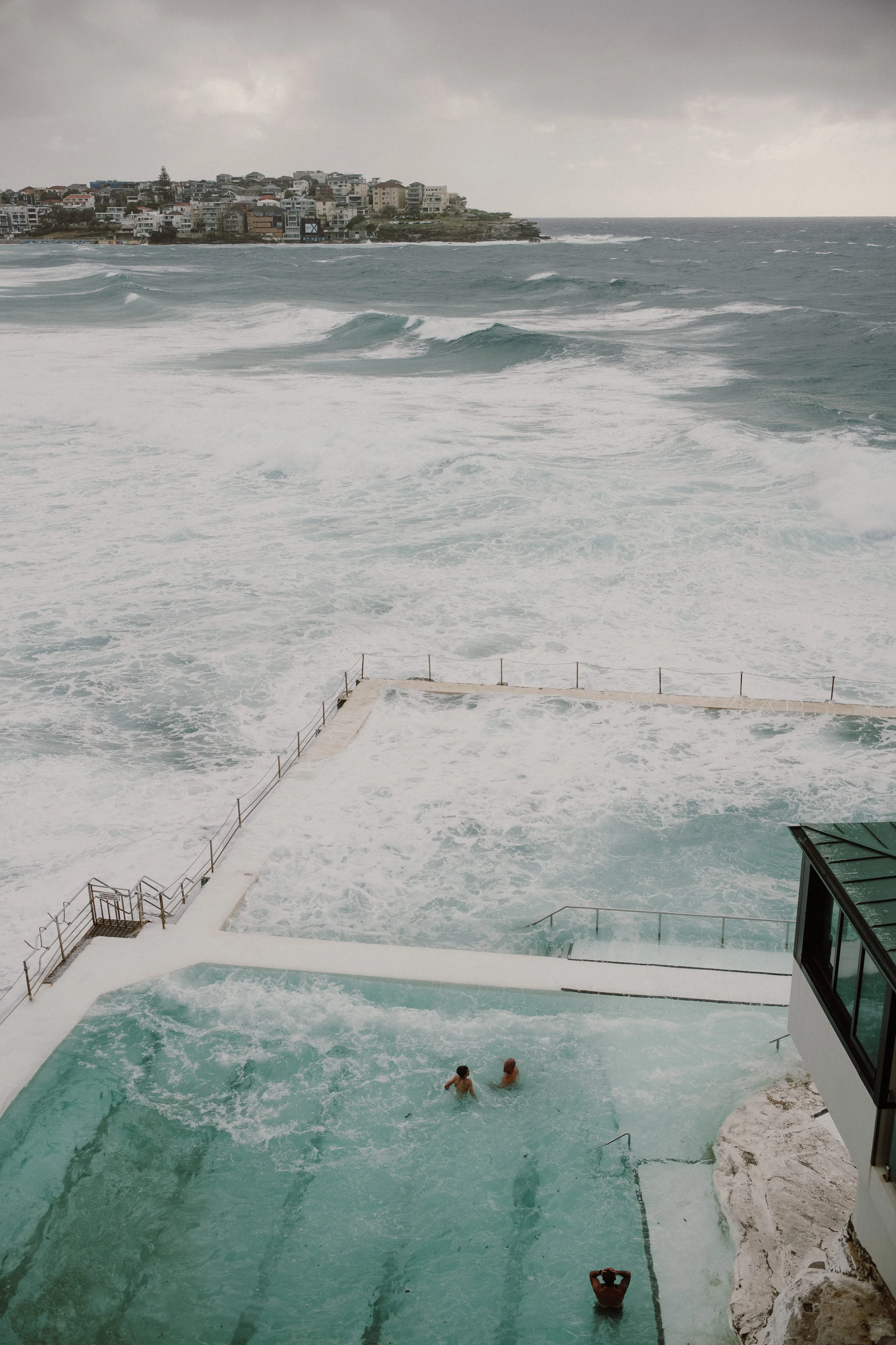 People swimming in an outdoor pool near the beach with large waves and overcast sky
