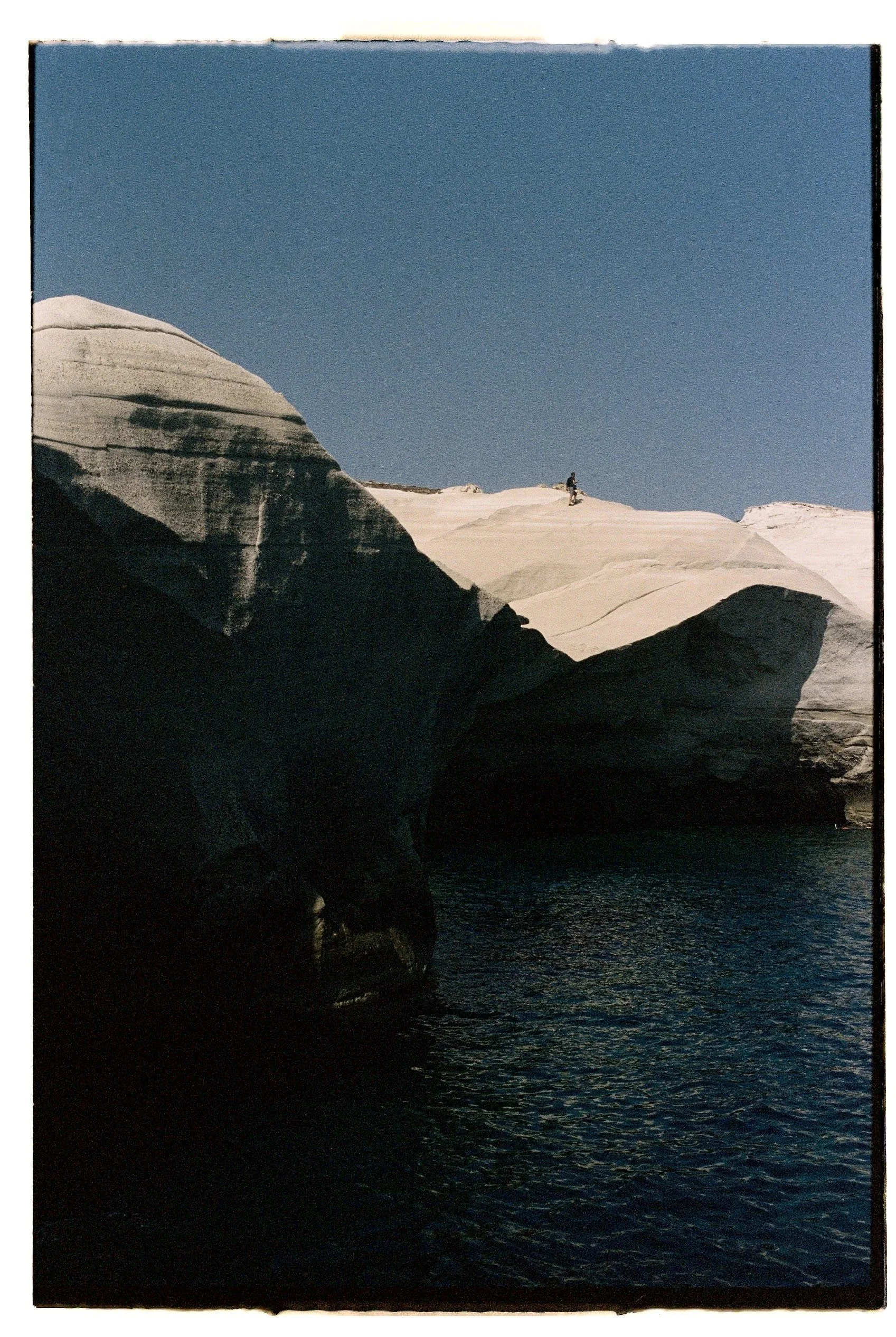 A person hiking on white, layered rocky cliffs above dark water with a clear blue sky overhead.