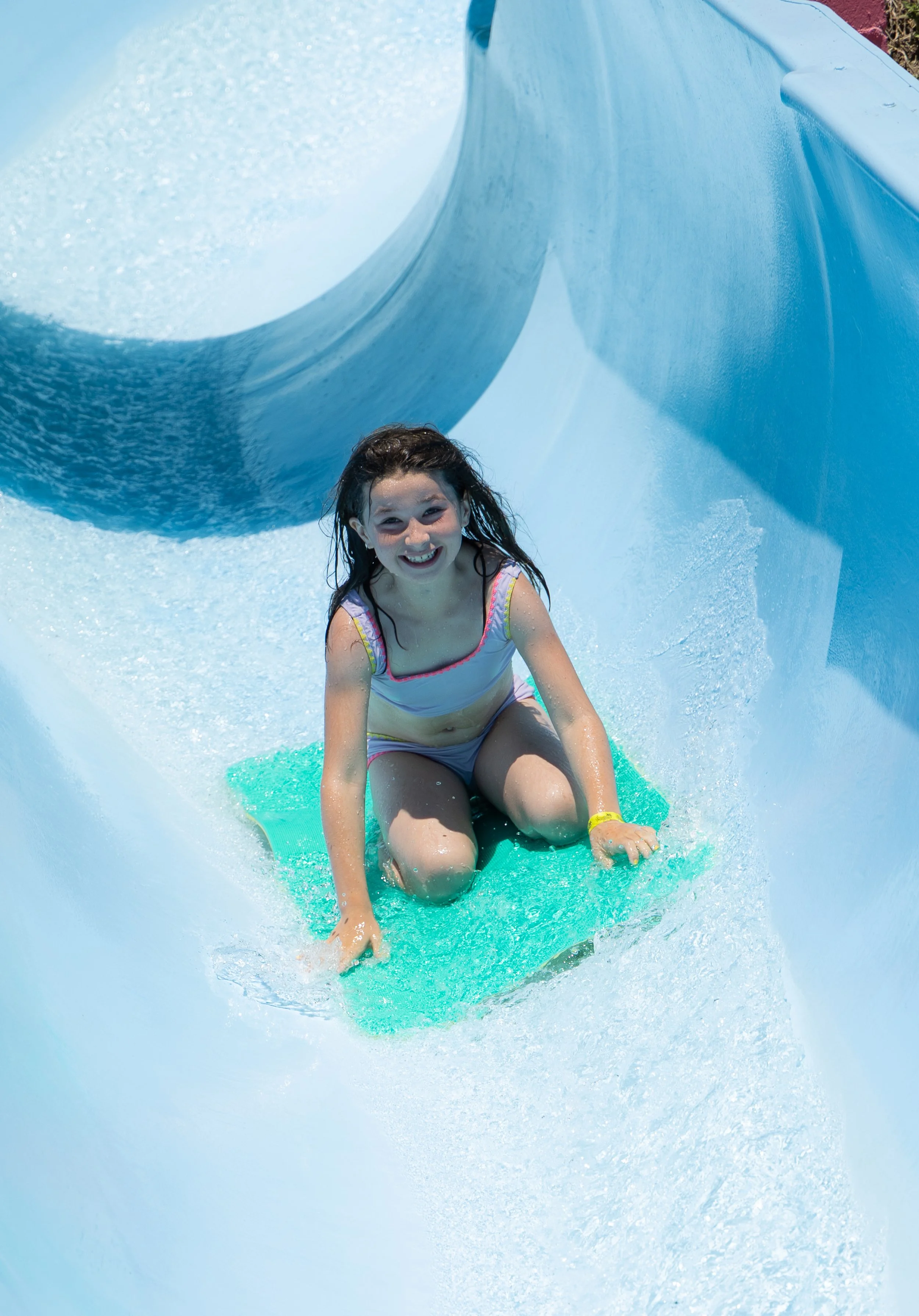Summer family fun at Mustang Waterpark in Arroyo Grande. A young girl with wet hair and a big smile riding a green mat in a water slide with blue sides, splashing water around her.