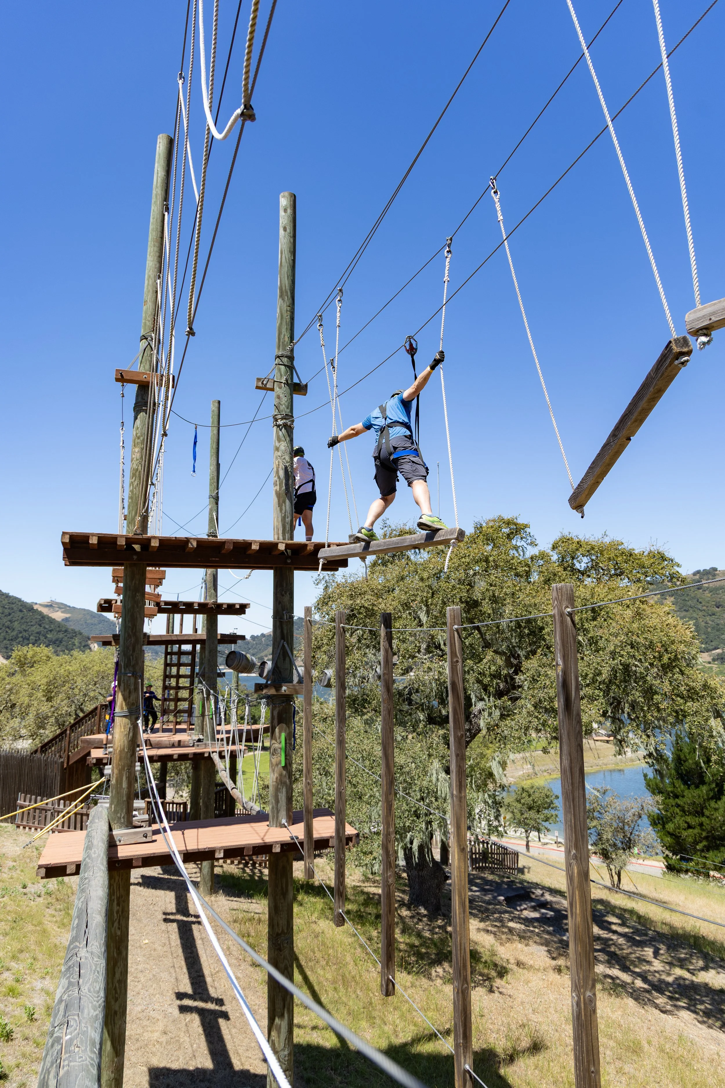 People navigating a ropes course with wooden and rope bridges outdoors on a sunny day, with trees and Lopez Lake in the background.