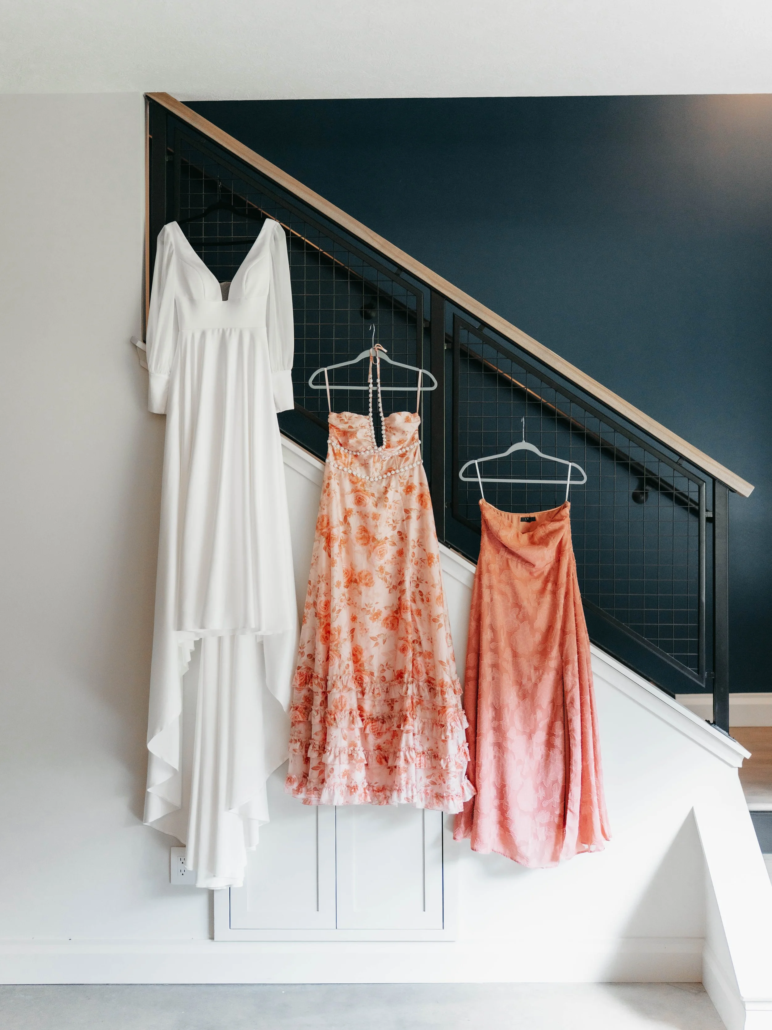 Three dresses hanging on a staircase railing, with a white gown on the left, a pink floral dress in the middle, and a peach-colored dress on the right.