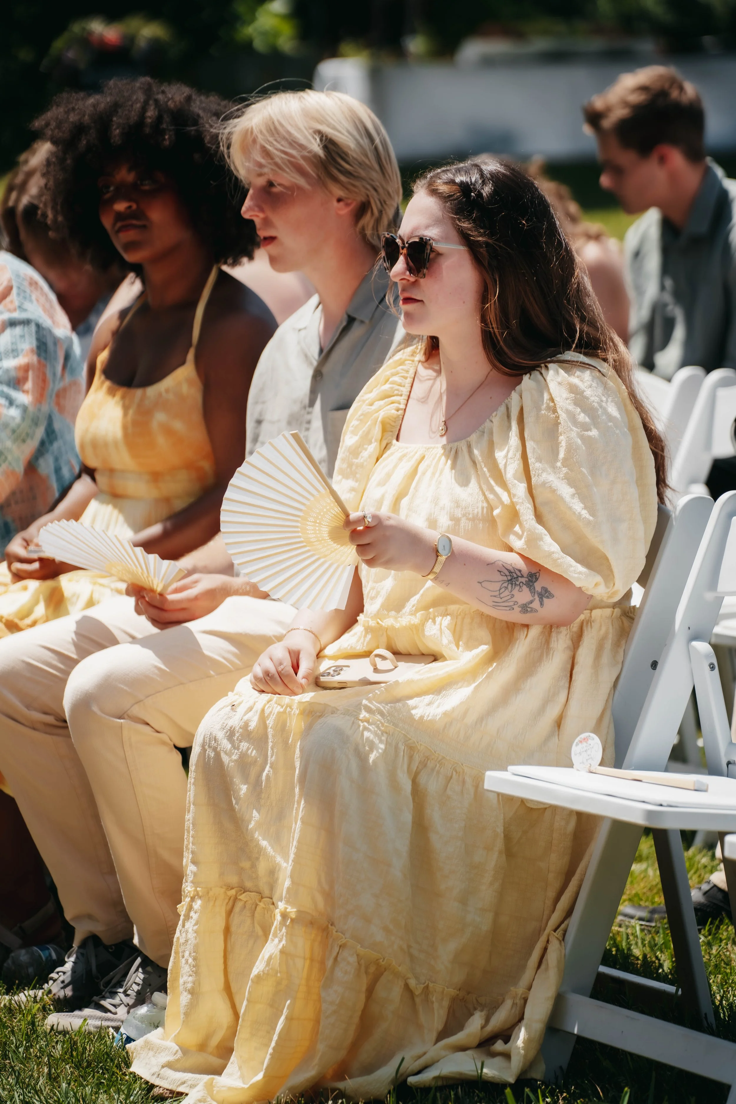 Group of people sitting in chairs outdoors during daytime, holding fans, dressed in light-colored clothing, engaging in a solemn event.