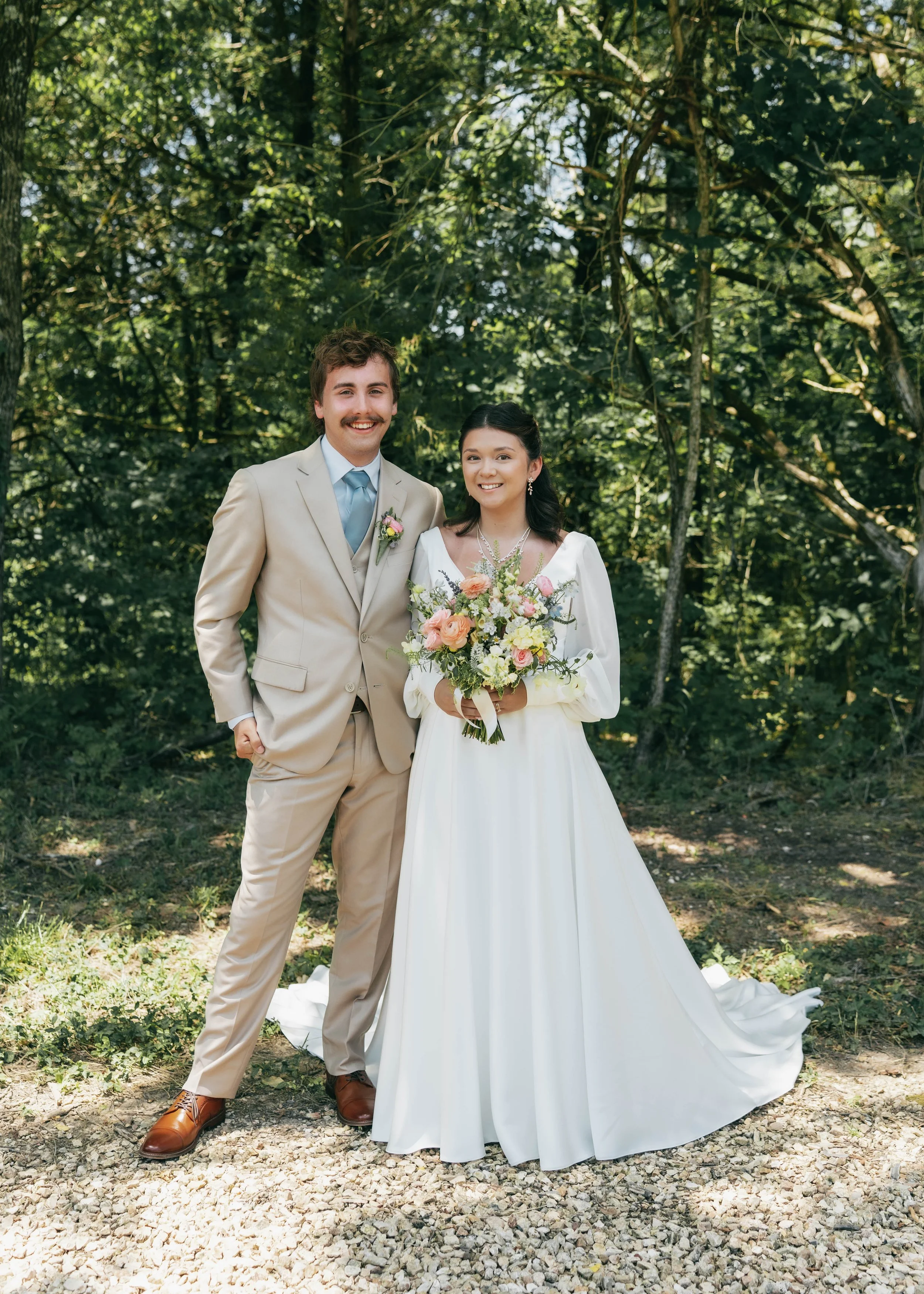 A bride and groom standing outdoors in a wooded area, smiling at the camera, with the bride holding a bouquet of pink and yellow flowers.