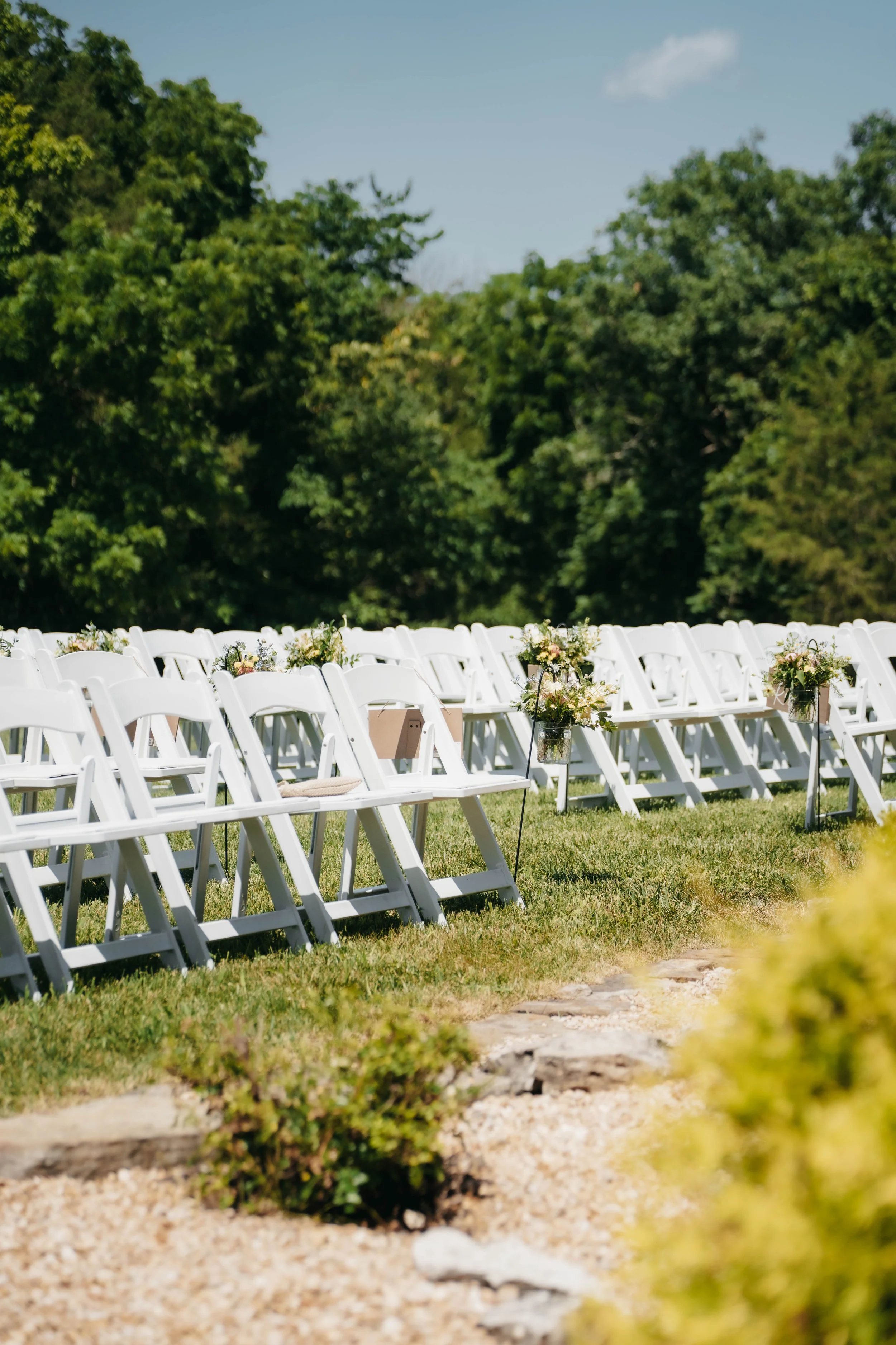 Empty white folding chairs decorated with floral arrangements set up outdoors on a grassy field for a wedding or event, with green trees and a blue sky in the background.