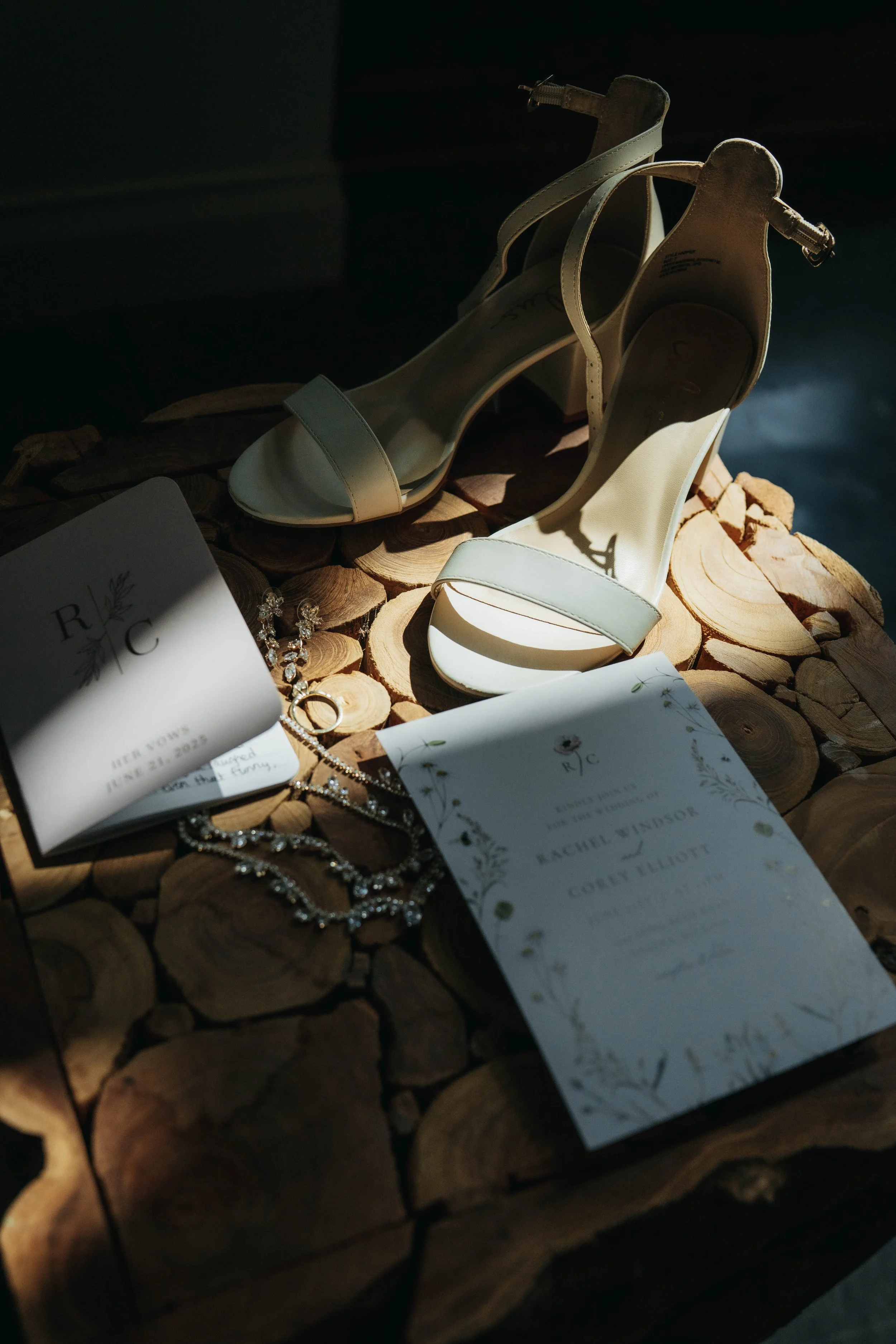 A pair of white high-heeled sandals, wedding invitation, and jewelry arranged on a wooden surface with sunlight shining on them.