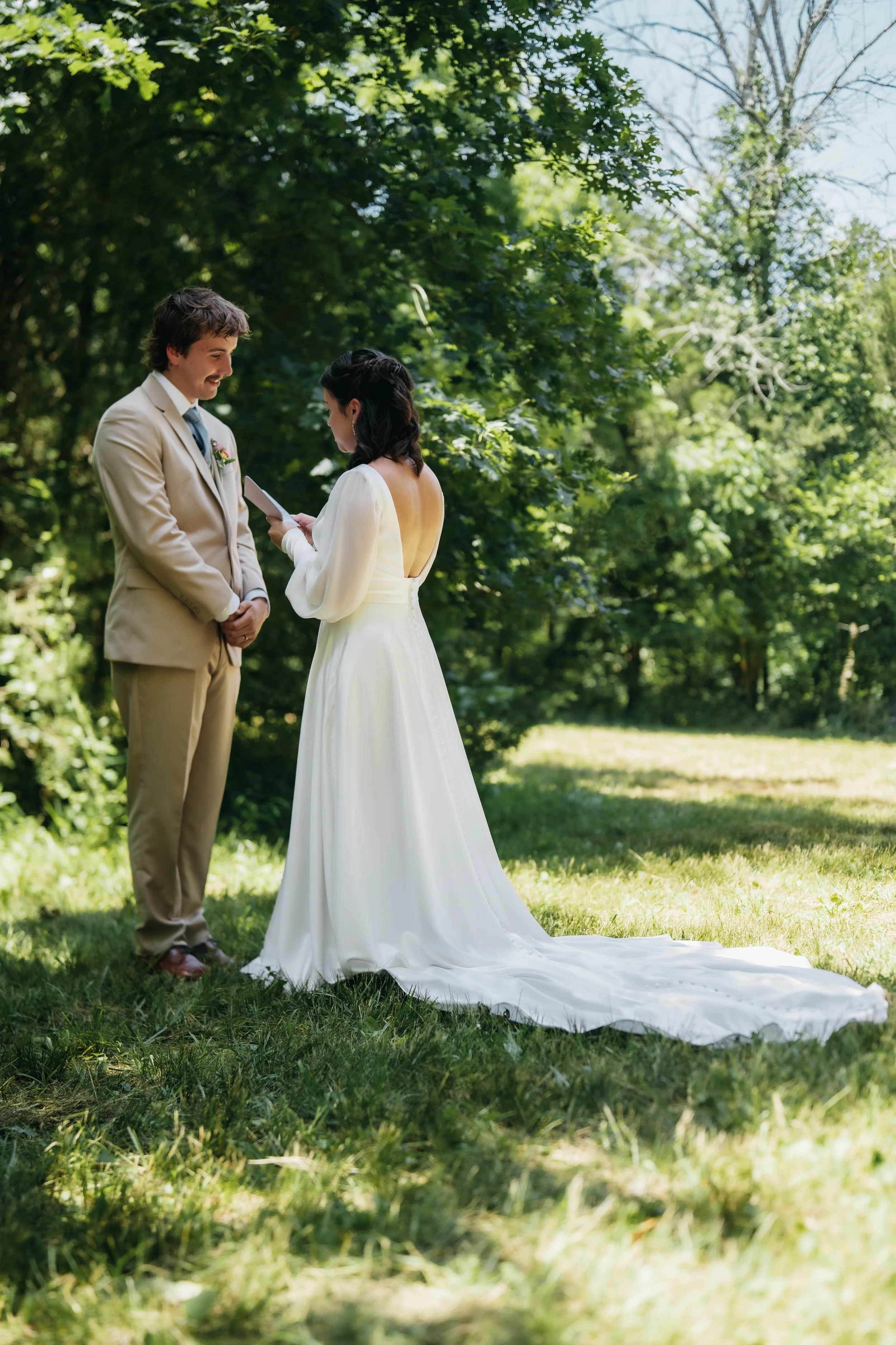 A bride and groom exchanging vows outdoors in a forest clearing, with greenery and trees in the background.