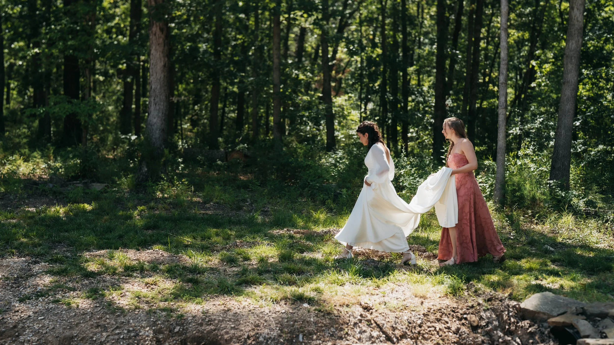 Two women in elegant dresses walking through a forest, one holding the train of the other's gown.