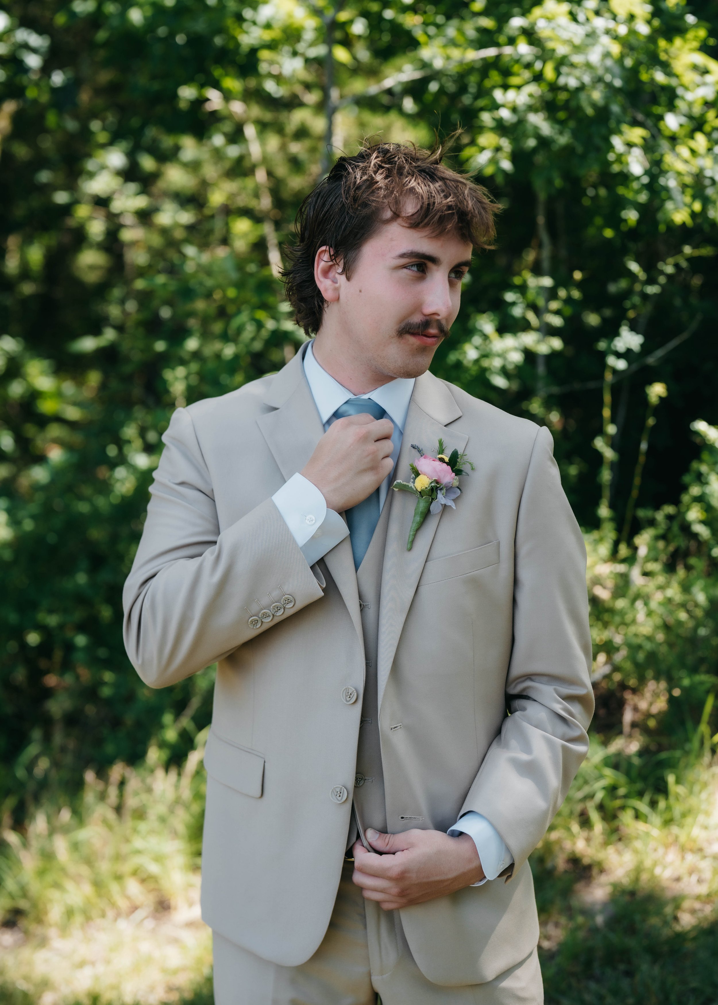 A groom dressed in a beige suit with a light blue tie and a boutonniere stands outdoors against a green, leafy background, adjusting his tie.