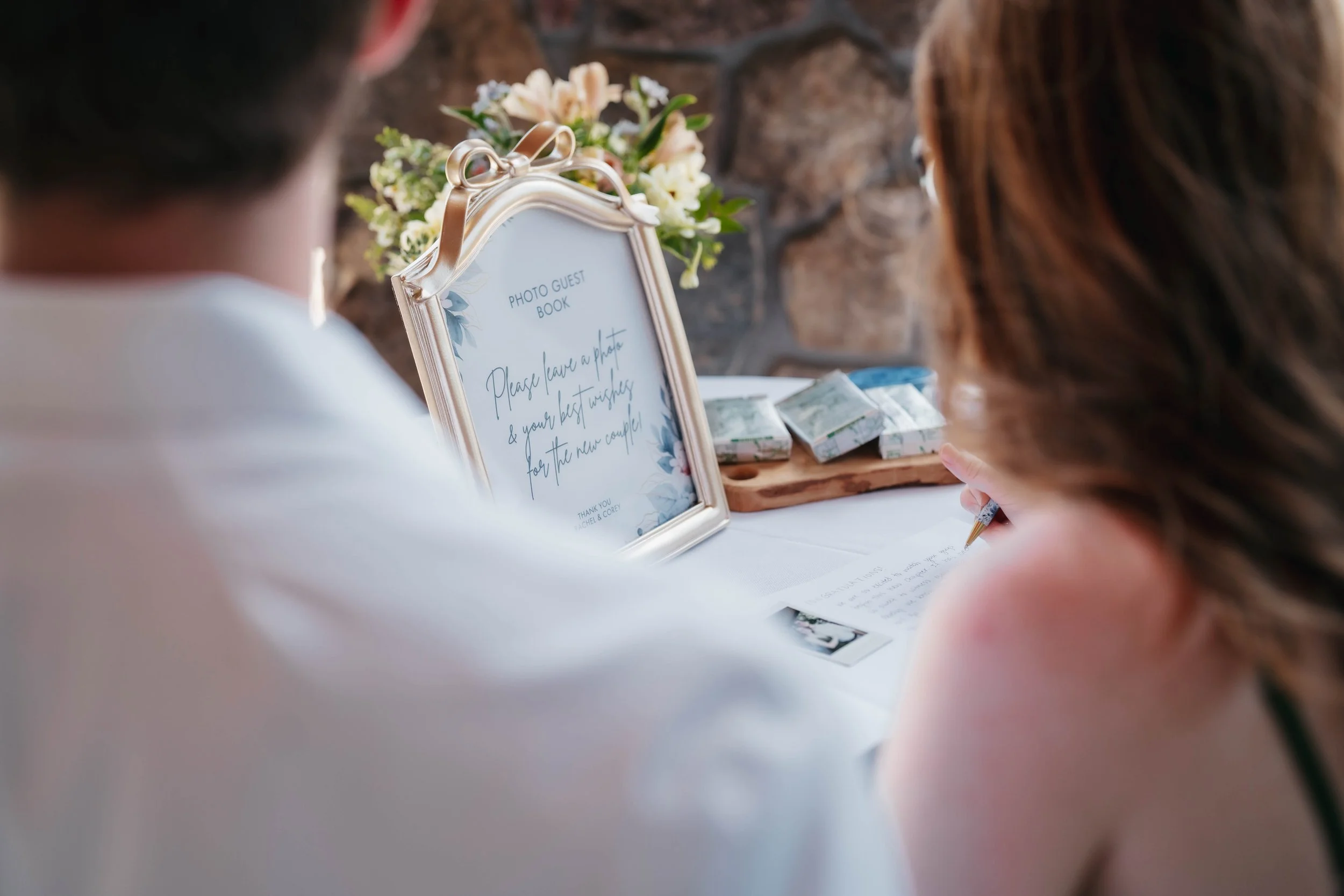 People at a wedding guest book table, with a frame sign that says, 'Please leave a photo & your best wishes for the new couple,' and a person writing a message on a card.