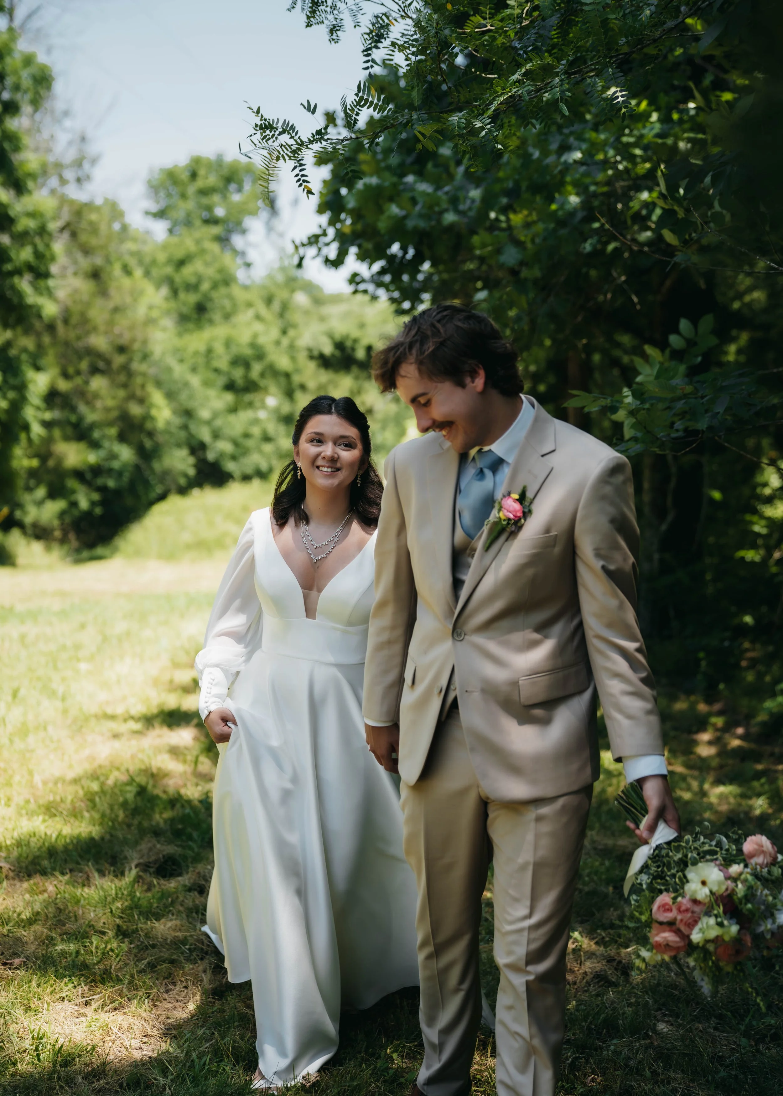 A bride in a white wedding gown and a groom in a beige suit walk in a green outdoor setting, smiling and holding flowers.