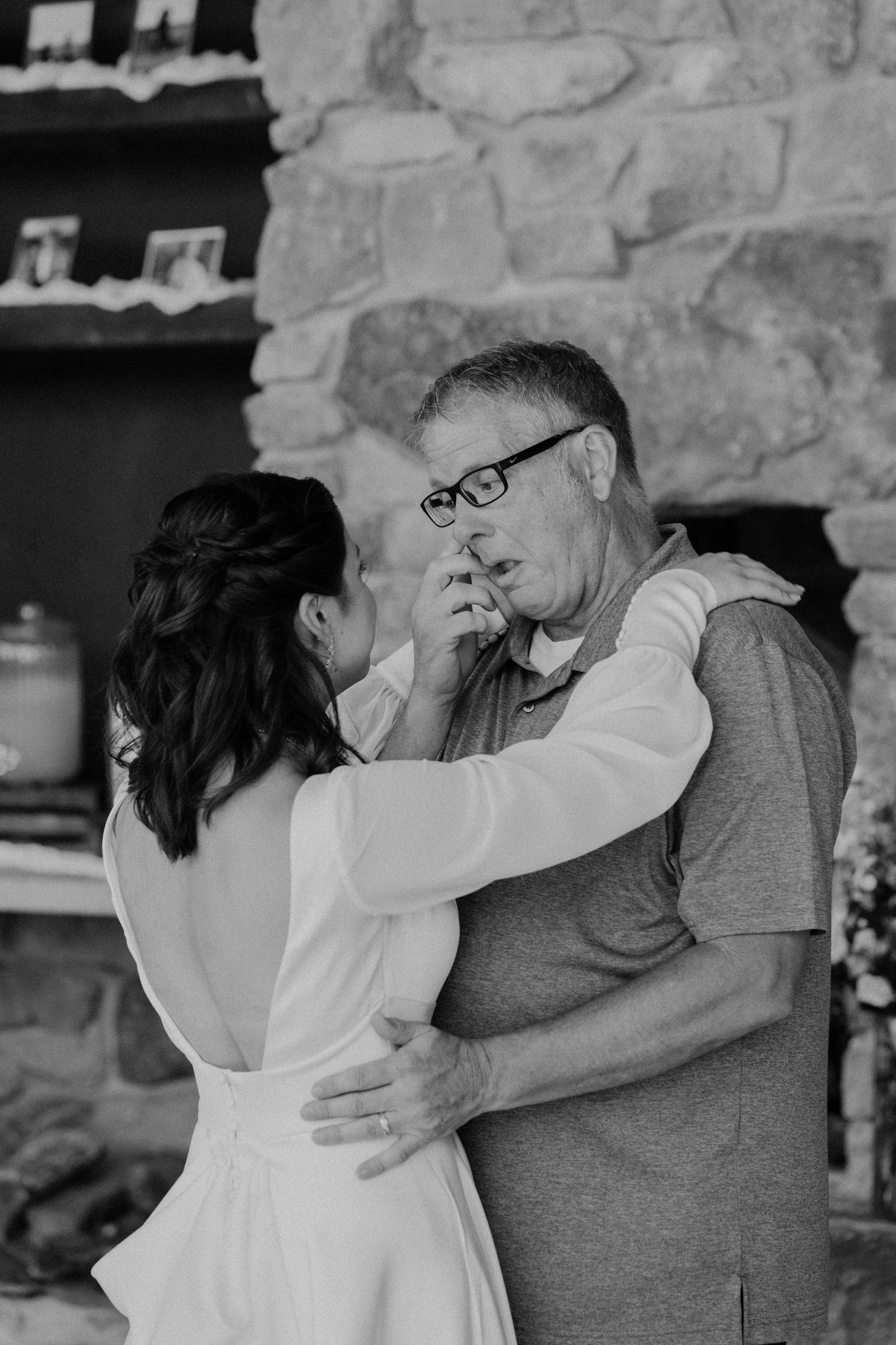 A woman in a white dress embraces an older man, touching his nose gently, in an emotional moment inside a rustic stone room.