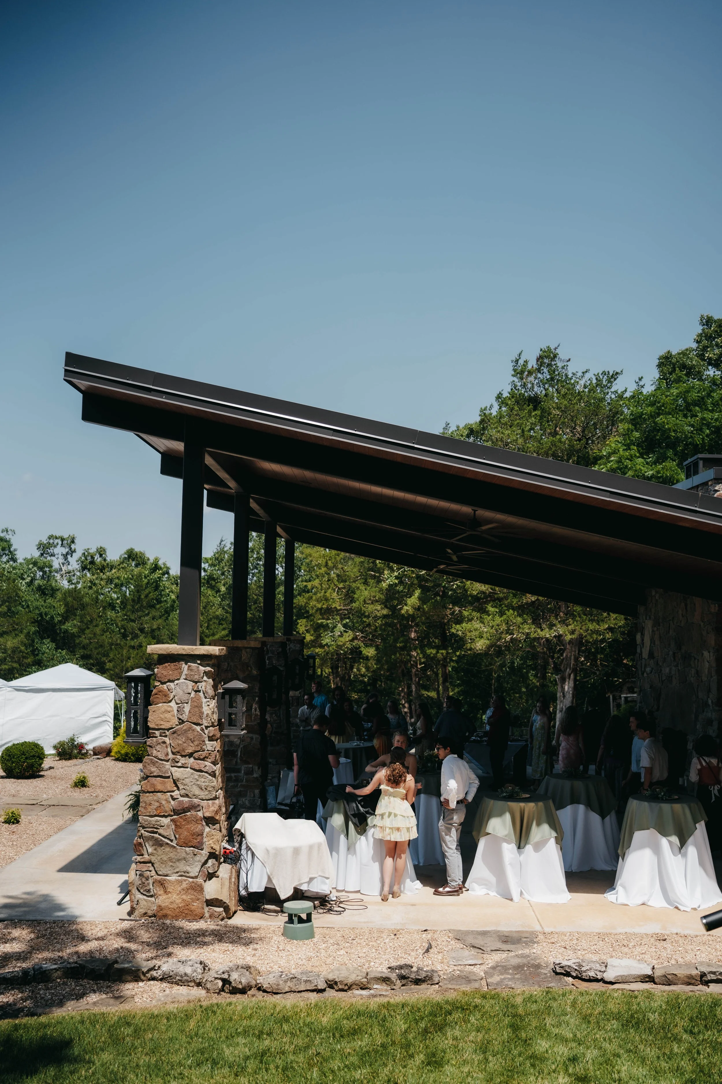 Outdoor gathering with people under a modern black-roofed pavilion with stone pillars, surrounded by greenery and a clear blue sky.