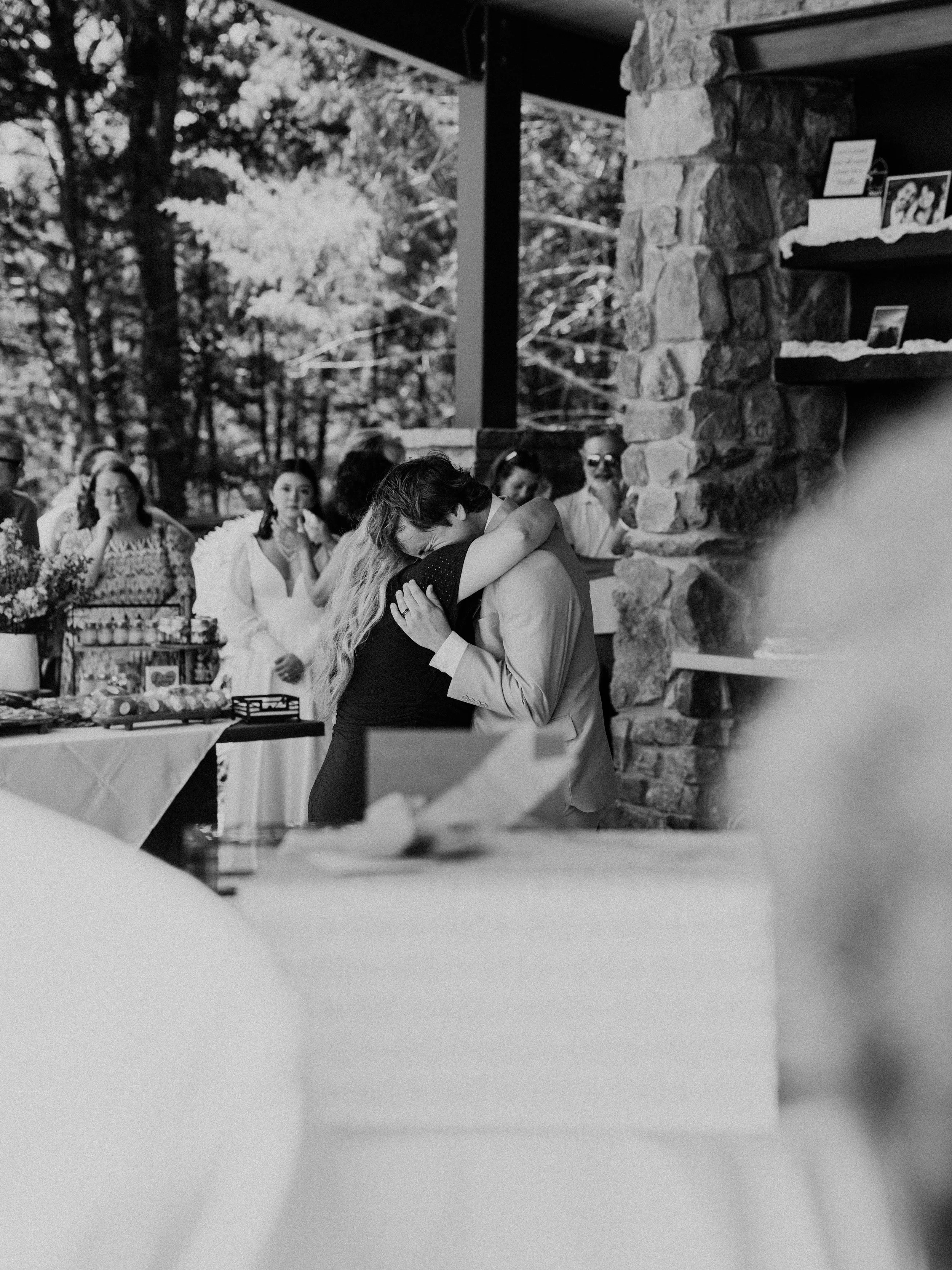 A black and white photo of a couple hugging each other in the center at a wedding reception, with guests in the background looking on and a table with desserts and decorations nearby.
