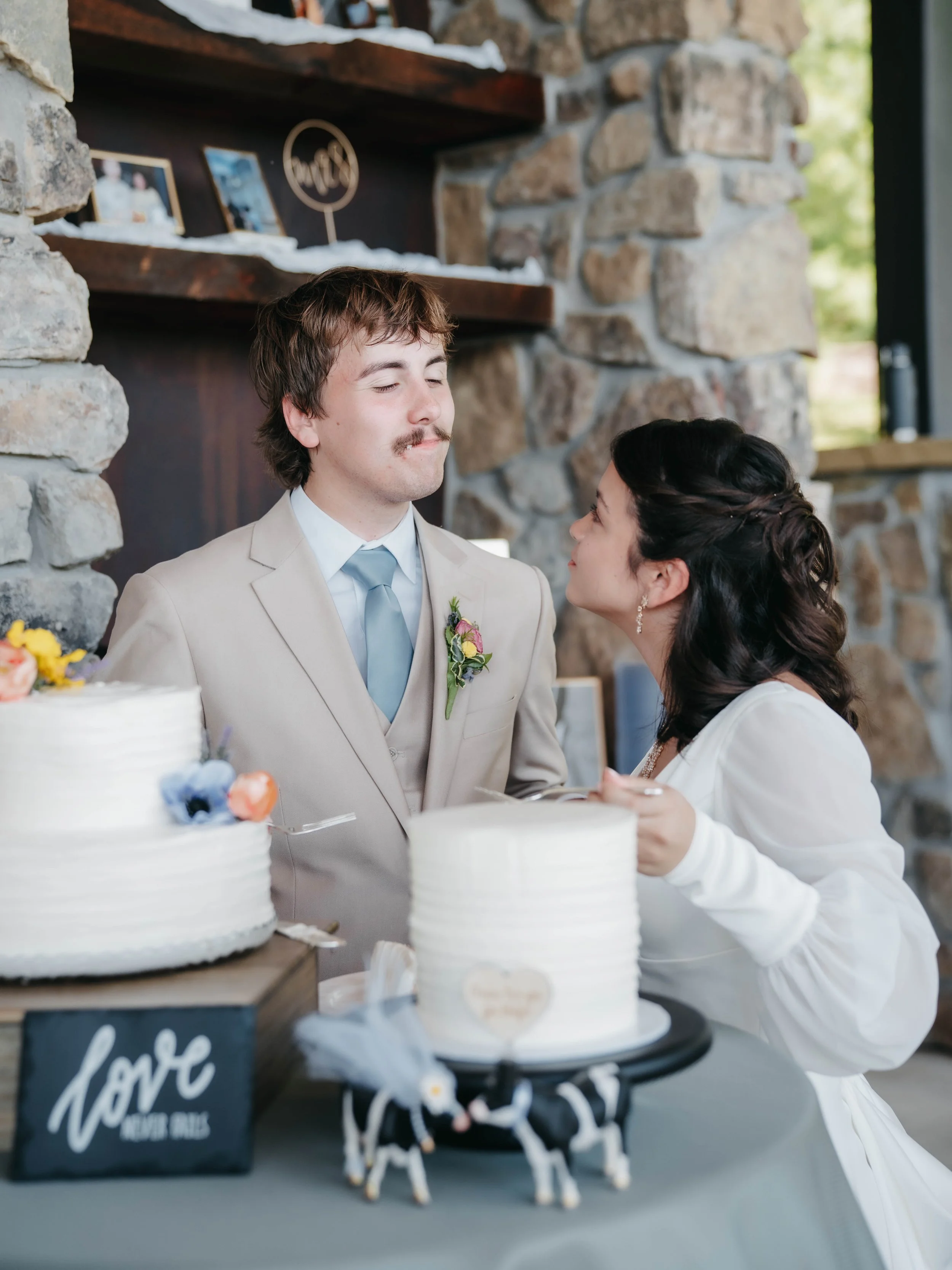 A bride and groom at their wedding reception, standing close together and smiling, with wedding cakes in front of them on a table decorated with a blackboard sign that says "love never fails" and cow figurines.