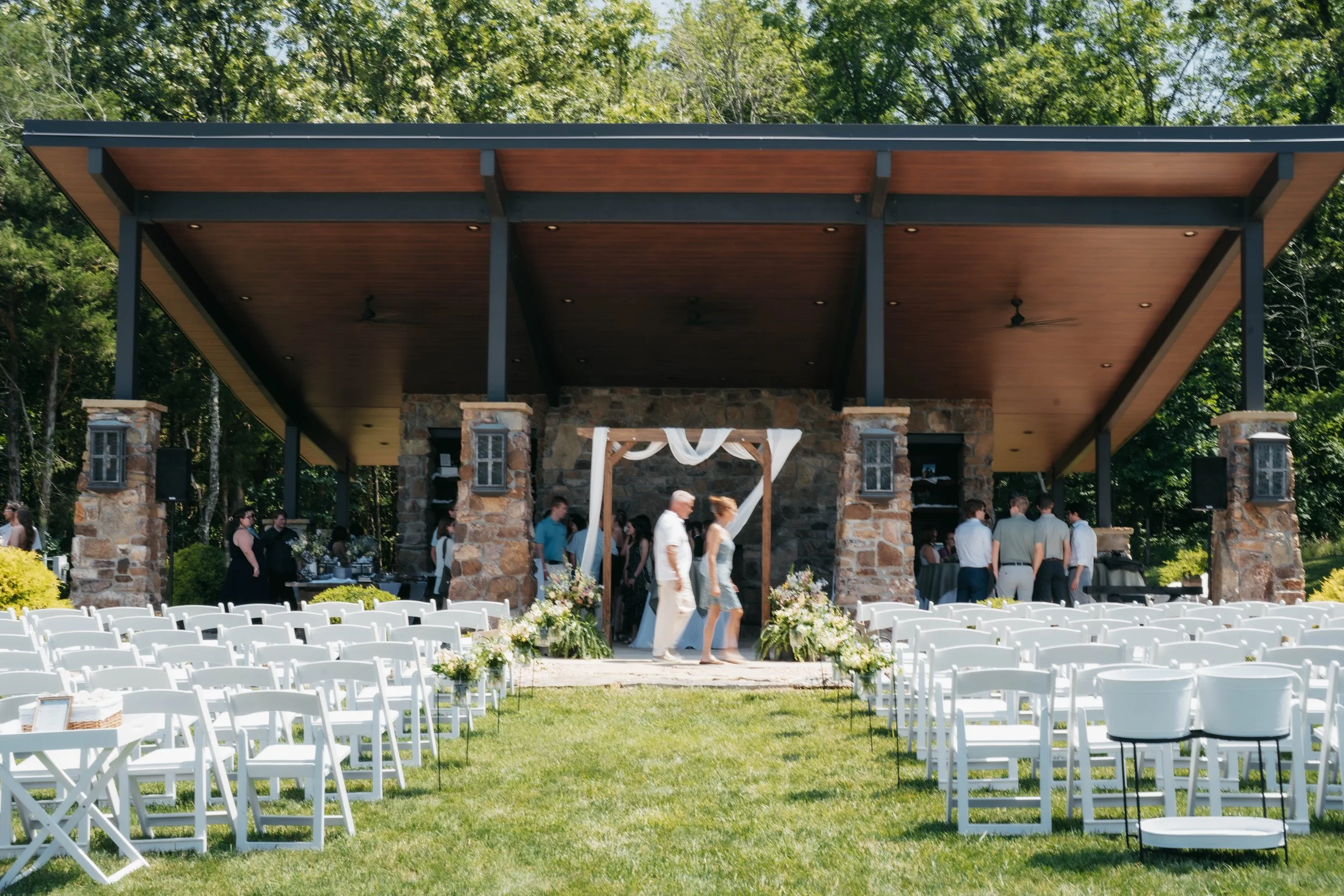 Outdoor wedding ceremony setup with white chairs aligned on a grassy area facing a stone and wood pavilion, decorated with floral arrangements, and guests gathering near the pavilion against a backdrop of trees.