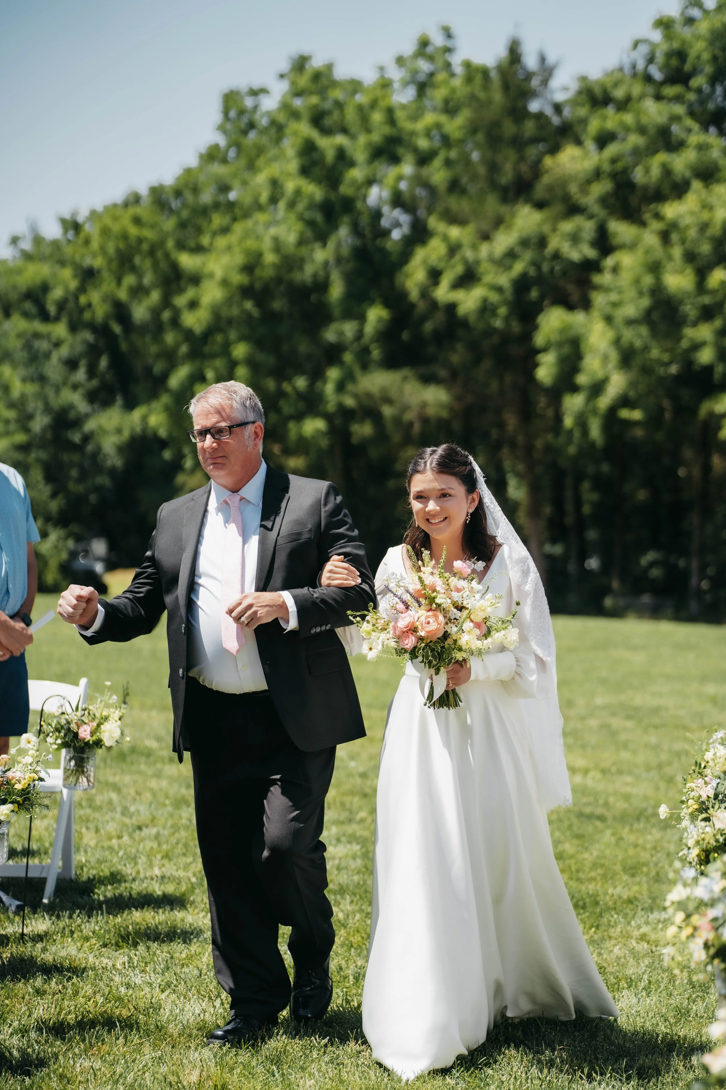 A bride in a white wedding dress holding a bouquet, walking outdoors with an older man in a black suit, on a grassy area with trees in the background.