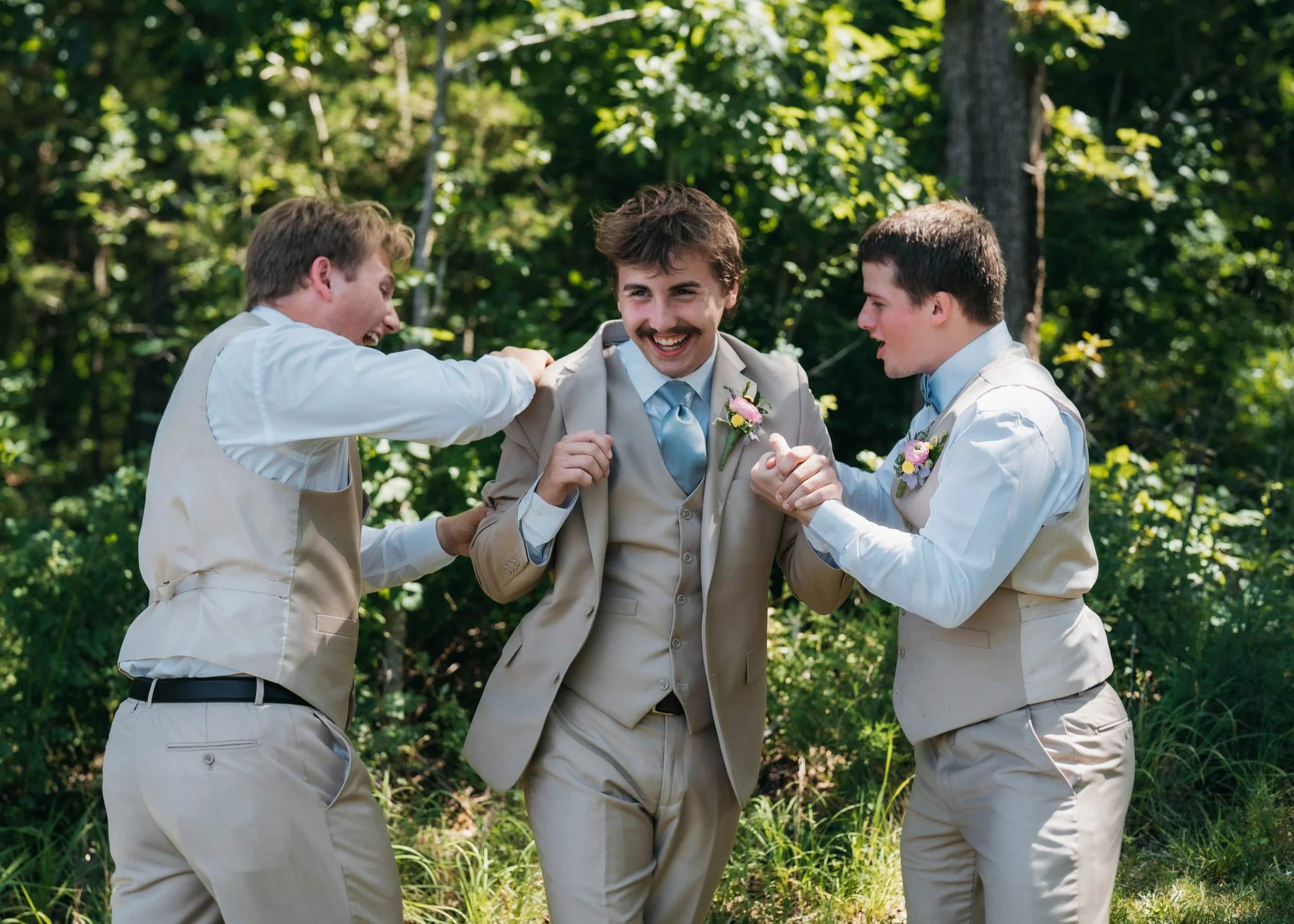 Three men dressed in beige suits and vests, standing outdoors in a wooded area, celebrating and smiling together.