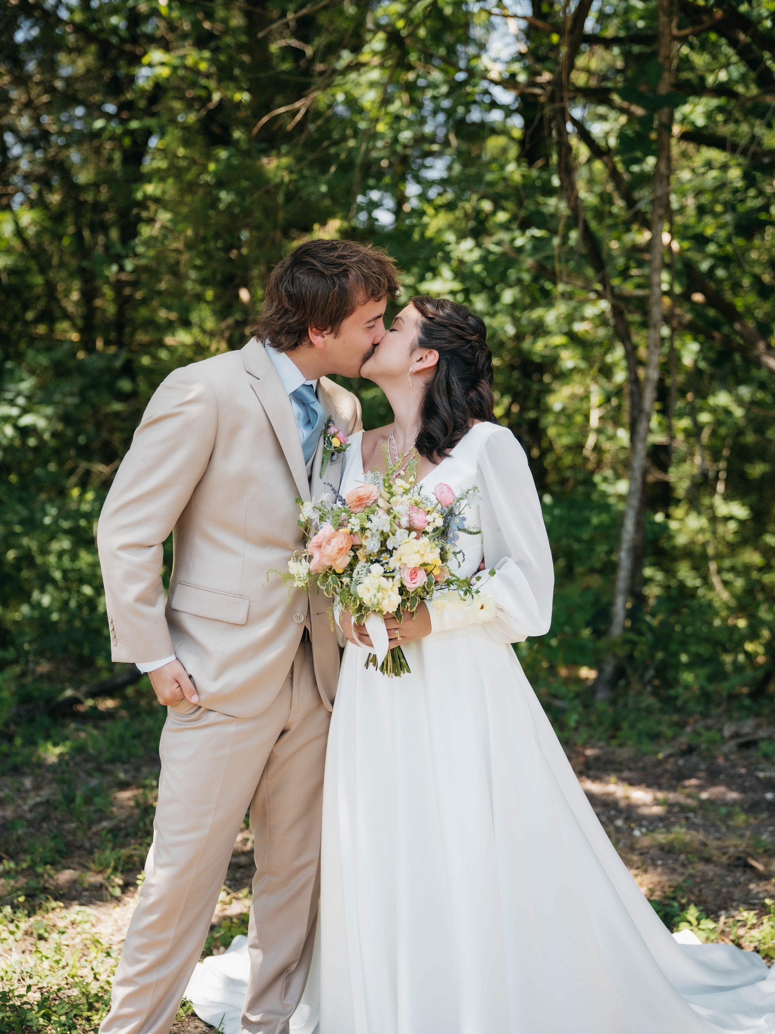 A bride and groom sharing a kiss outdoors during their wedding in a lush green wooded area. The bride is holding a bouquet of flowers and wearing a long white dress, while the groom is dressed in a light beige suit.