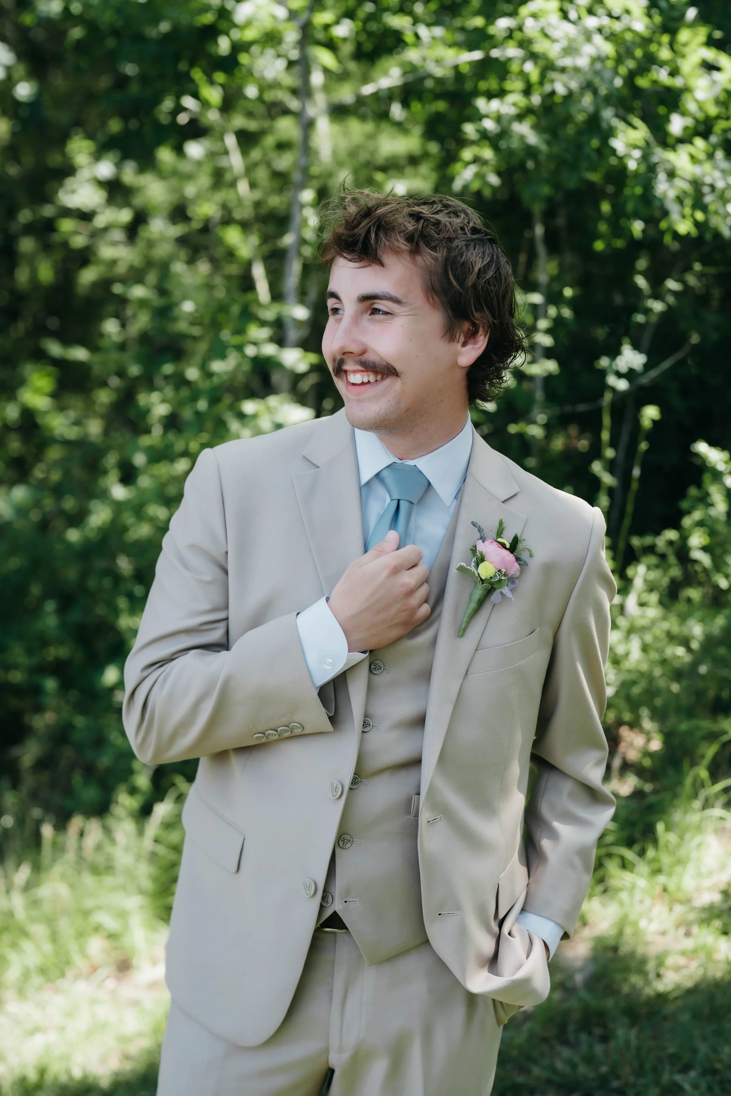 A young man in a beige suit with a light blue shirt and tie, smiling outdoors with greenery in the background, wearing a boutonnière.
