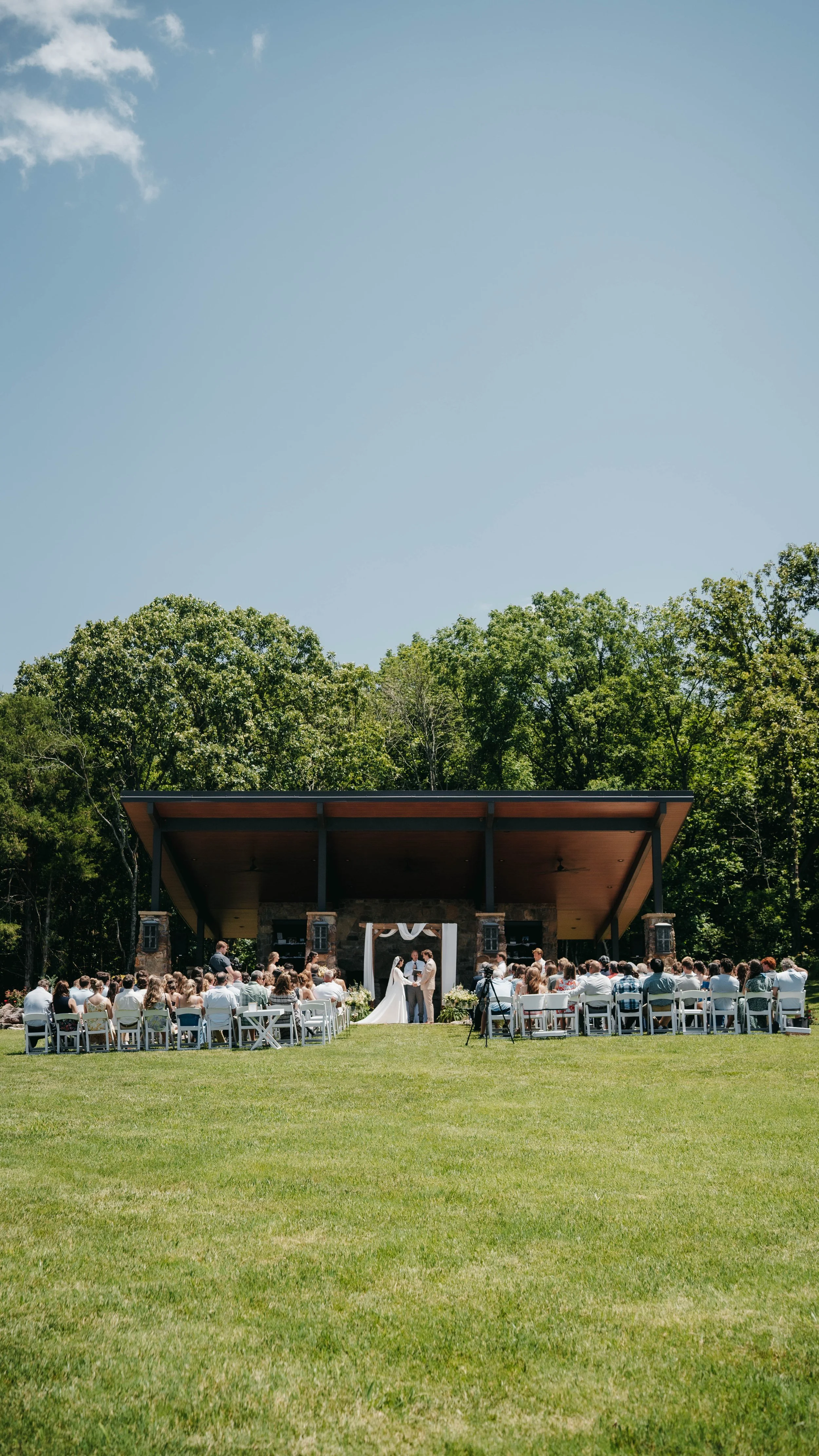 Outdoor wedding ceremony on a grass field with a wooden stage, officiant, and bride and groom, surrounded by seated guests, with a backdrop of trees and a clear blue sky.
