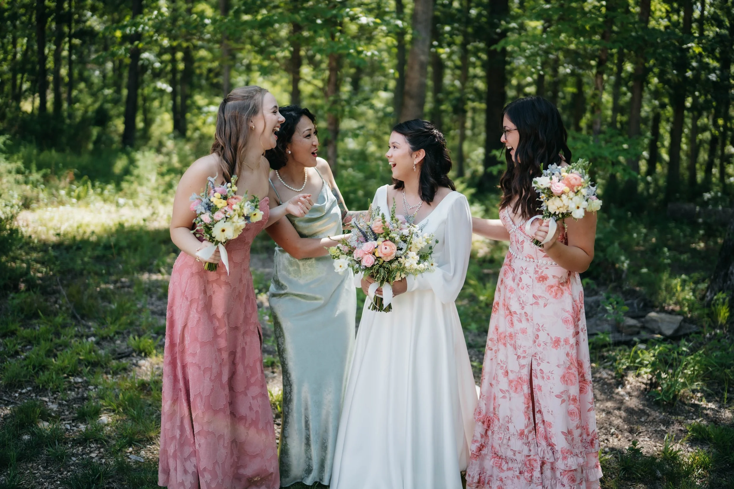 Four women dressed in elegant dresses, holding bouquets, smiling and celebrating outdoors in a forest setting.
