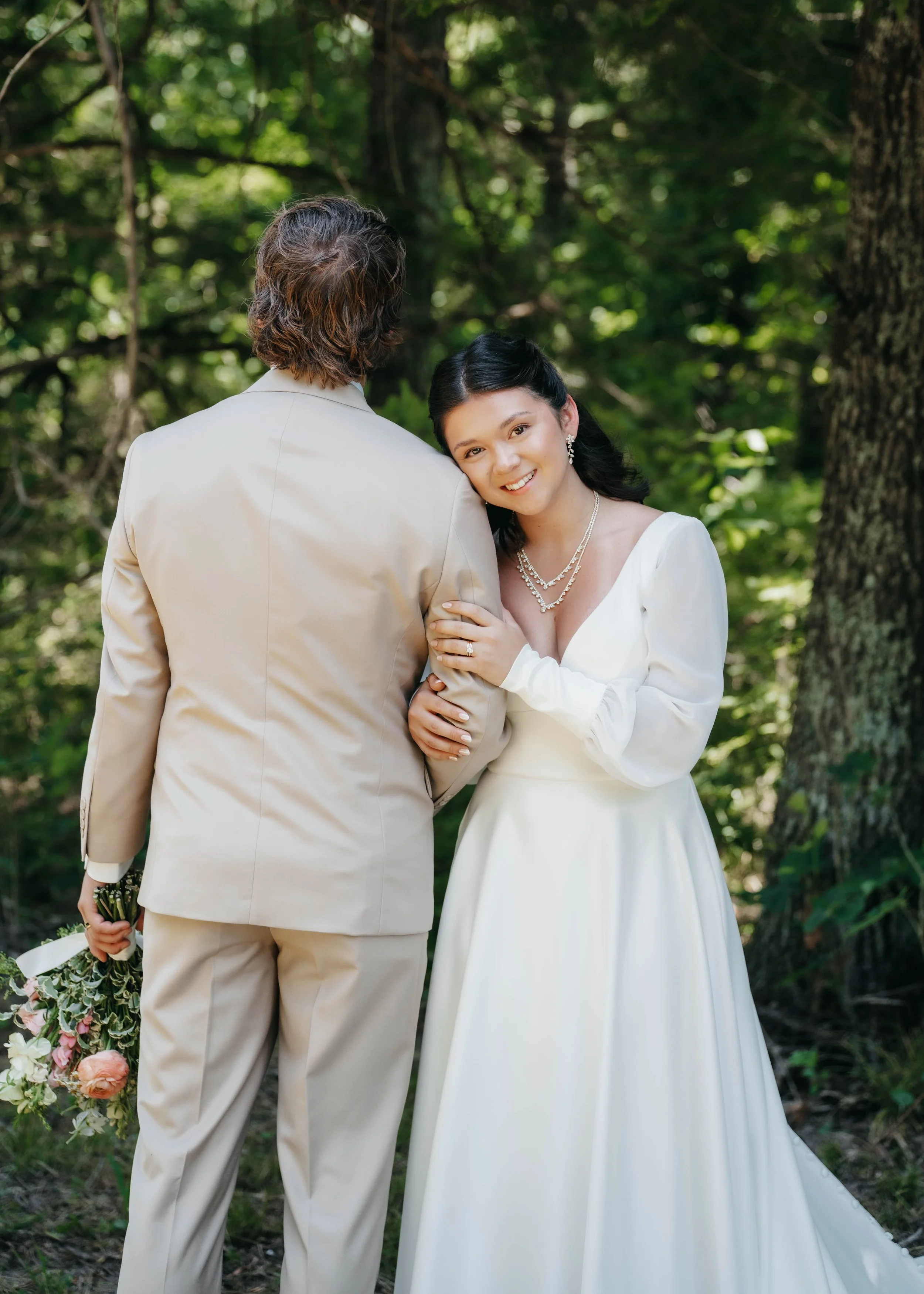 A bride and groom embrace outdoors in a forested area, with the bride smiling at the camera. She is wearing a white wedding dress and jewelry, while the groom is dressed in a beige suit and holding a bouquet of flowers.