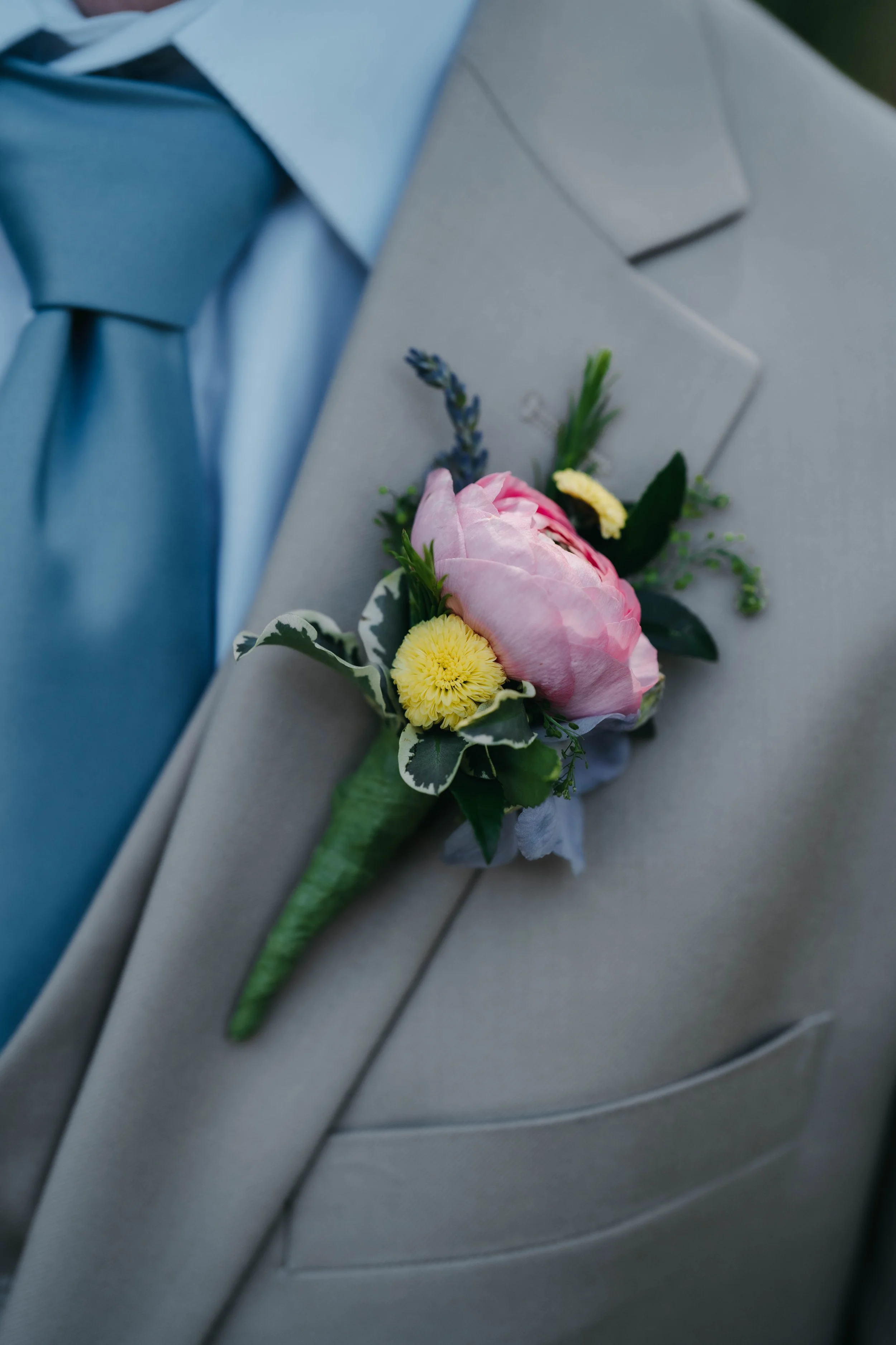 Close-up of a man's gray suit with a pink flower boutonniere pinned to the lapel, featuring a pink peony, yellow daisy, lavender sprig, greenery, and variegated leaves, with a blue shirt and matching blue tie.