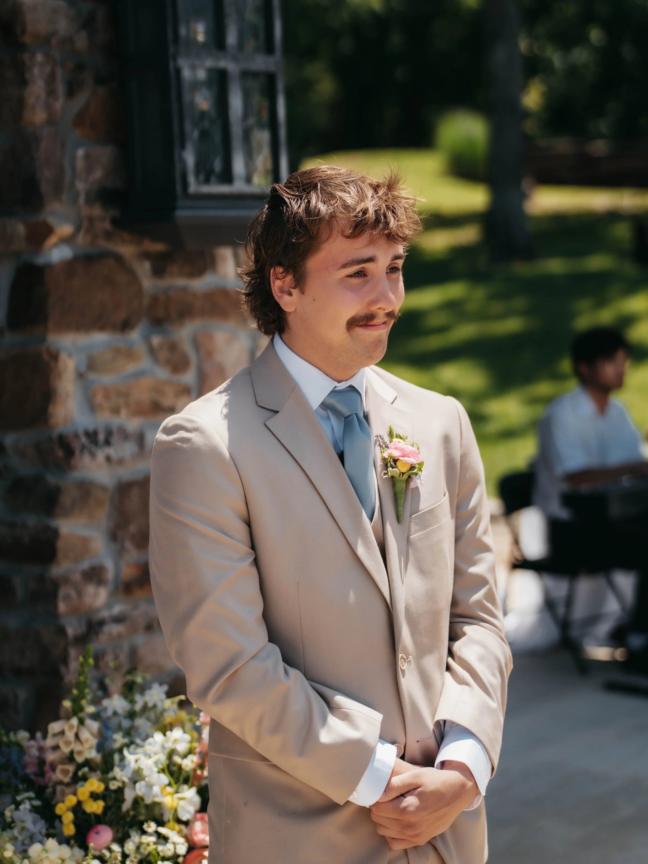 A man with a brown mustache and wavy hair dressed in a light beige suit with a white shirt, light blue tie, and a pink flower boutonniere, standing outdoors with a thoughtful expression.
