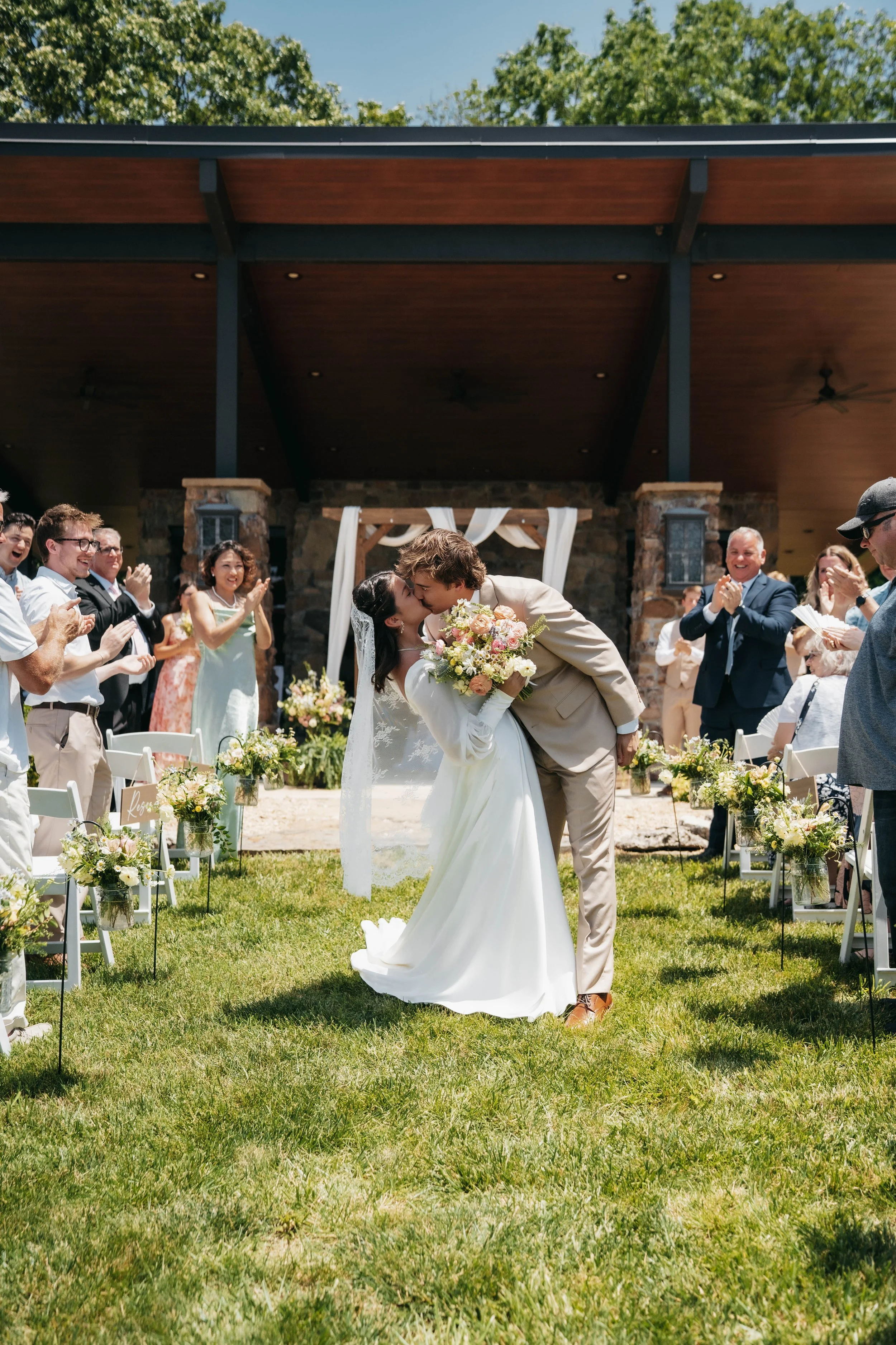 Bride and groom sharing a kiss during their outdoor wedding ceremony with guests clapping and celebrating.