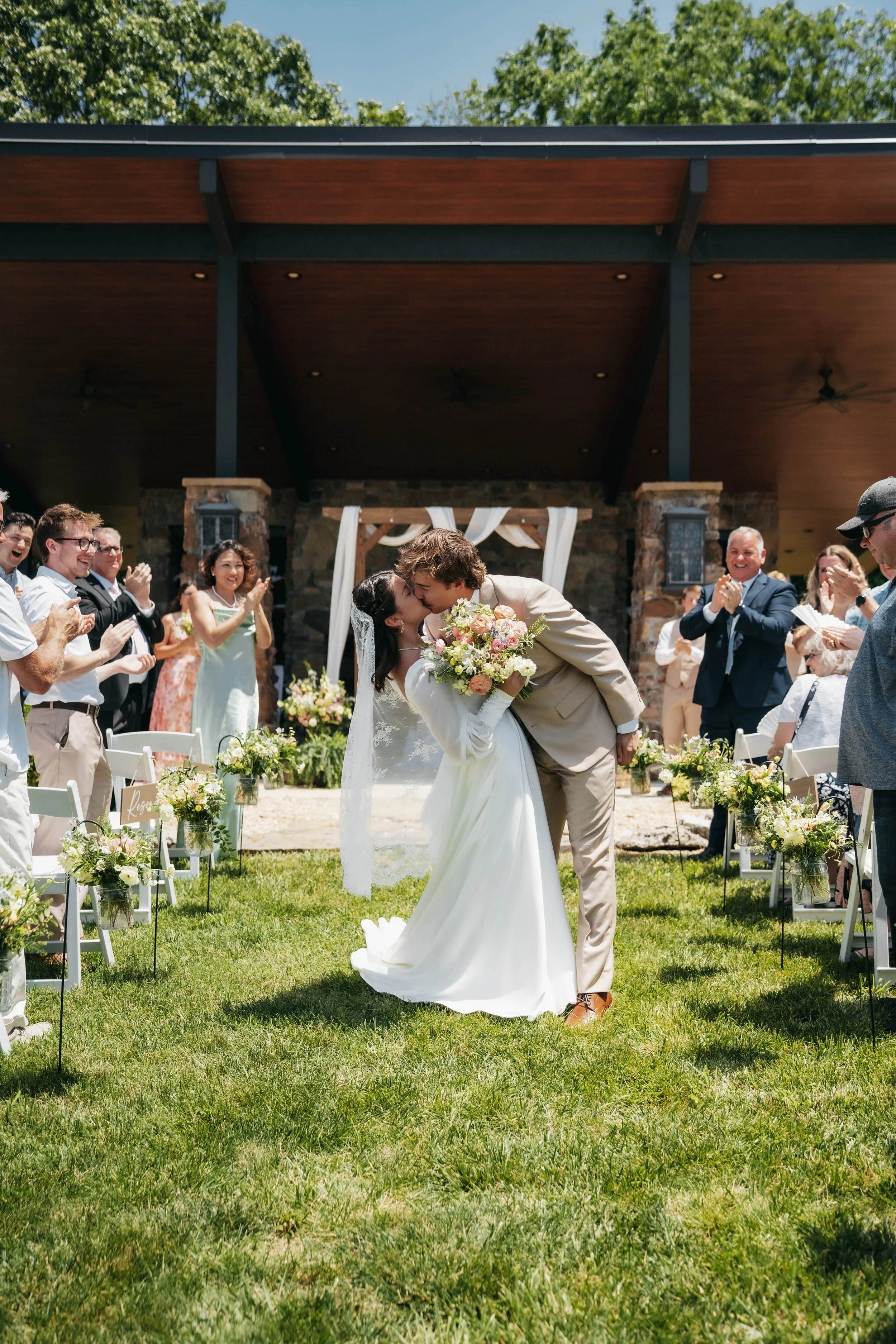 Bride and groom sharing a kiss during their outdoor wedding ceremony, surrounded by guests clapping and smiling, with floral decorations and a wooden pavilion in the background.