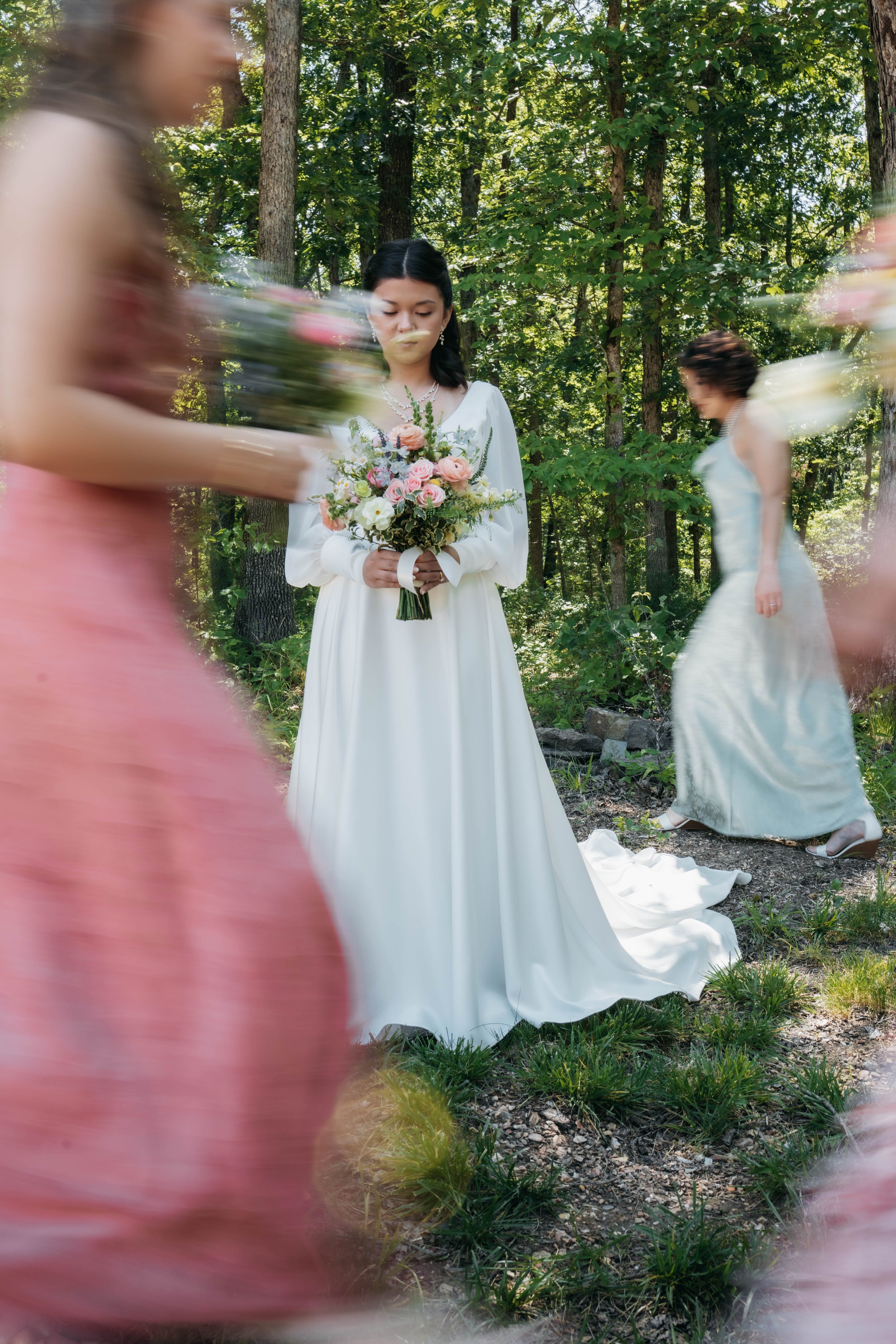 A bride in a white wedding dress holding a bouquet of pink and white flowers, standing in a forest surrounded by blurred women in pastel-colored dresses, with sunlight filtering through trees.